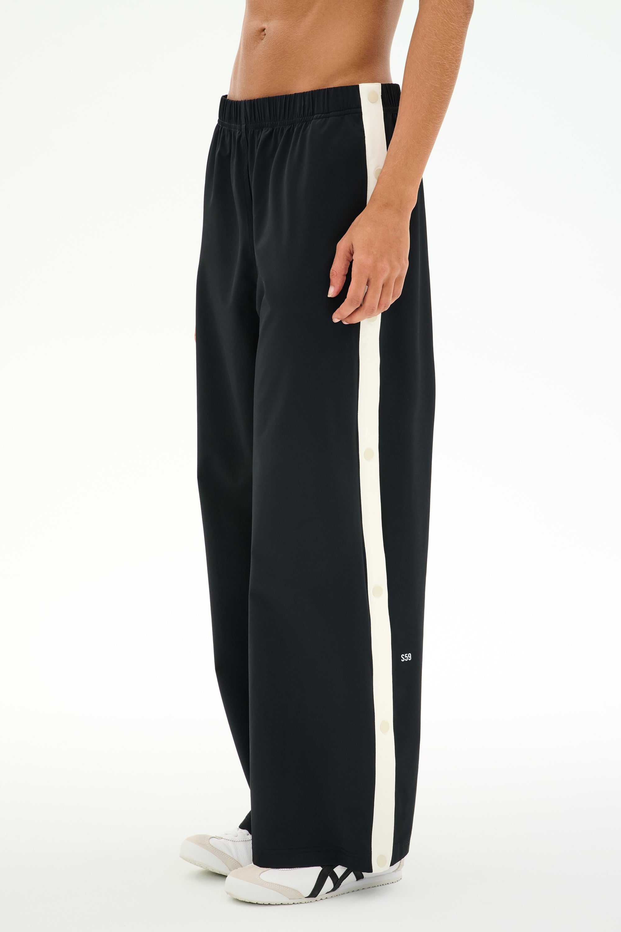 A person wears the SPLITS59 Jordan Nylon Twill Tearaway pants in Black/Creme, featuring a comfortable nylon-spandex blend, wide-leg fit, white side stripe, and snap buttons, paired with white sneakers.