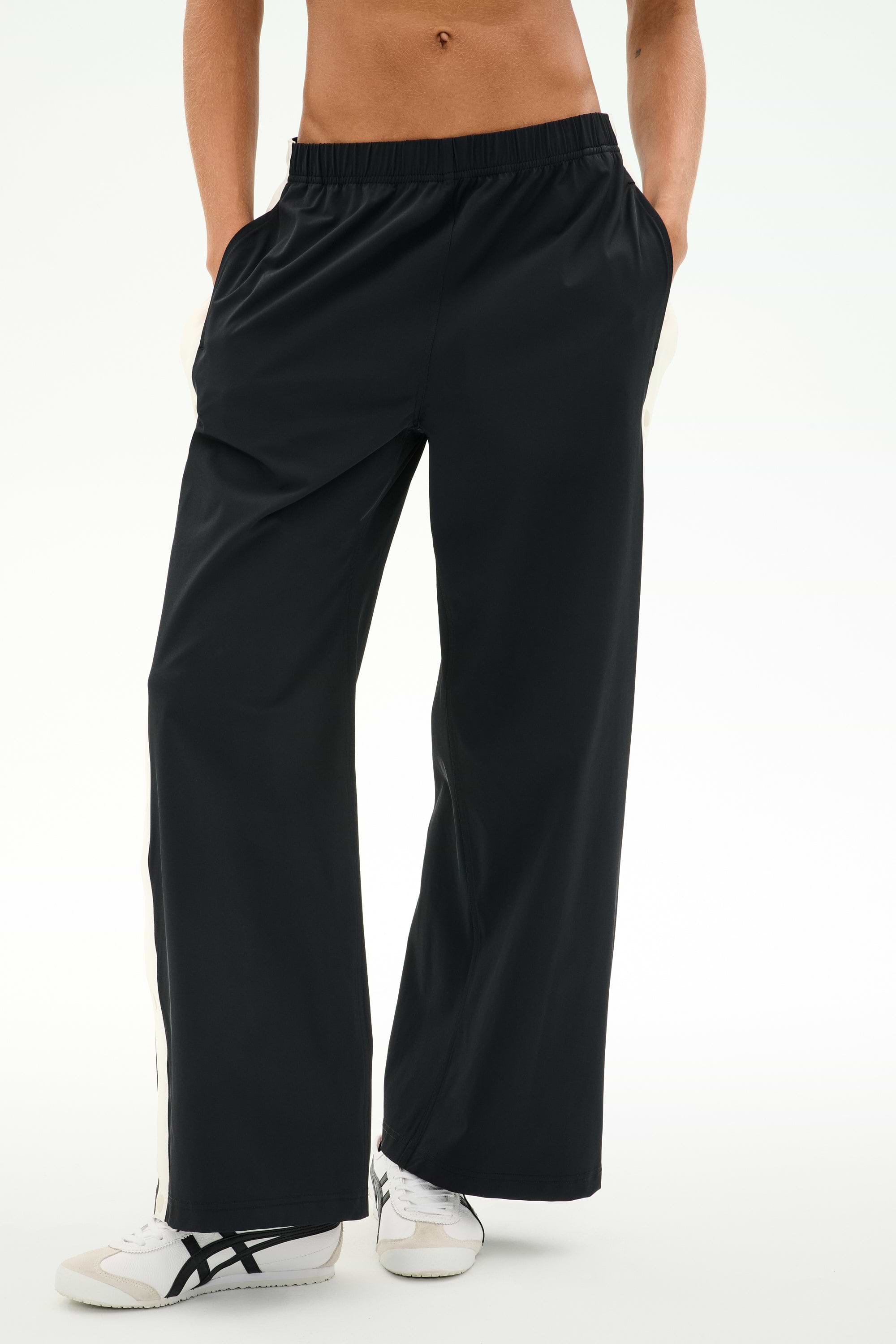 A person wearing the SPLITS59 Jordan Nylon Twill Tearaway track pants in Black/Creme, crafted from a lightweight nylon-spandex blend, stands with hands in pockets and white sneakers with black stripes against a plain background.