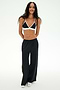 A person stands and smiles against a plain white background, wearing SPLITS59's Jordan Nylon Twill Tearaway pants in Black/Creme, paired with a black and white spandex bikini-style top and white shoes.