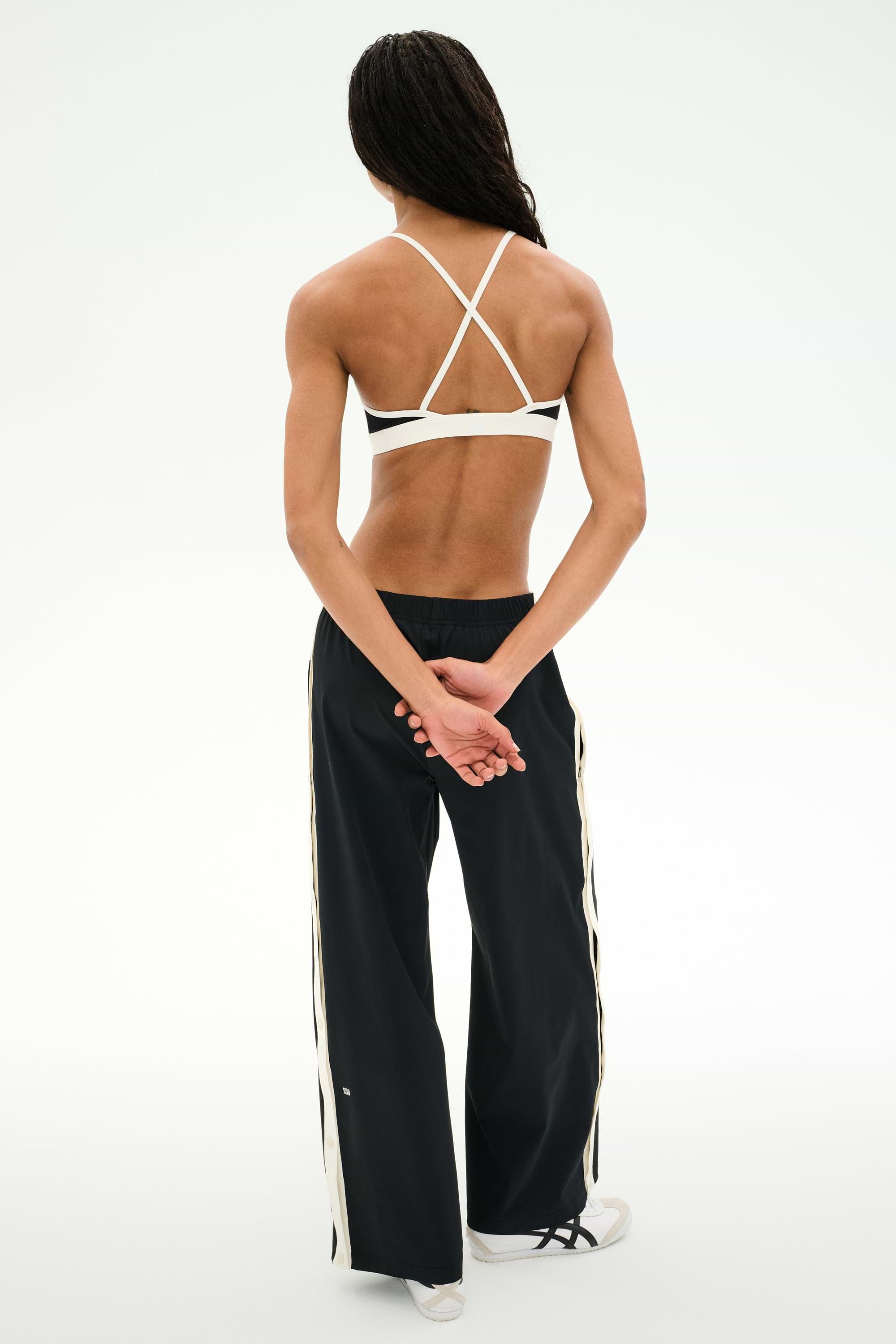 A person stands with hands behind their back, facing away from the camera against a plain background, wearing black SPLITS59 Jordan Nylon Twill Tearaway pants in Black/Creme featuring white stripes, paired with a white strappy sports bra and white sneakers.