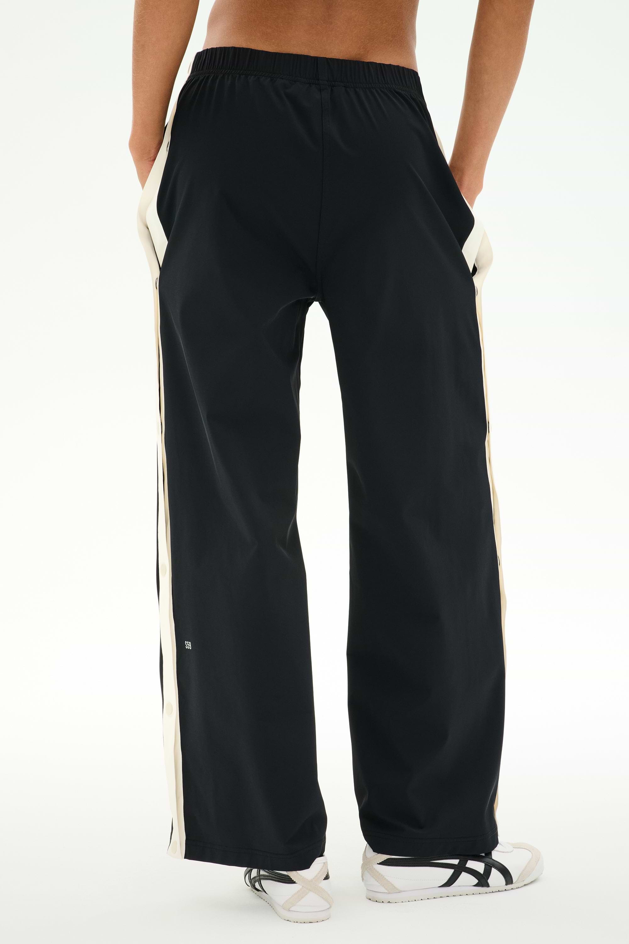 A person is seen from behind with hands in pockets, wearing SPLITS59's Jordan Nylon Twill Tearaway pants in Black/Creme, featuring white side stripes and crafted from a comfortable blend of nylon and spandex.
