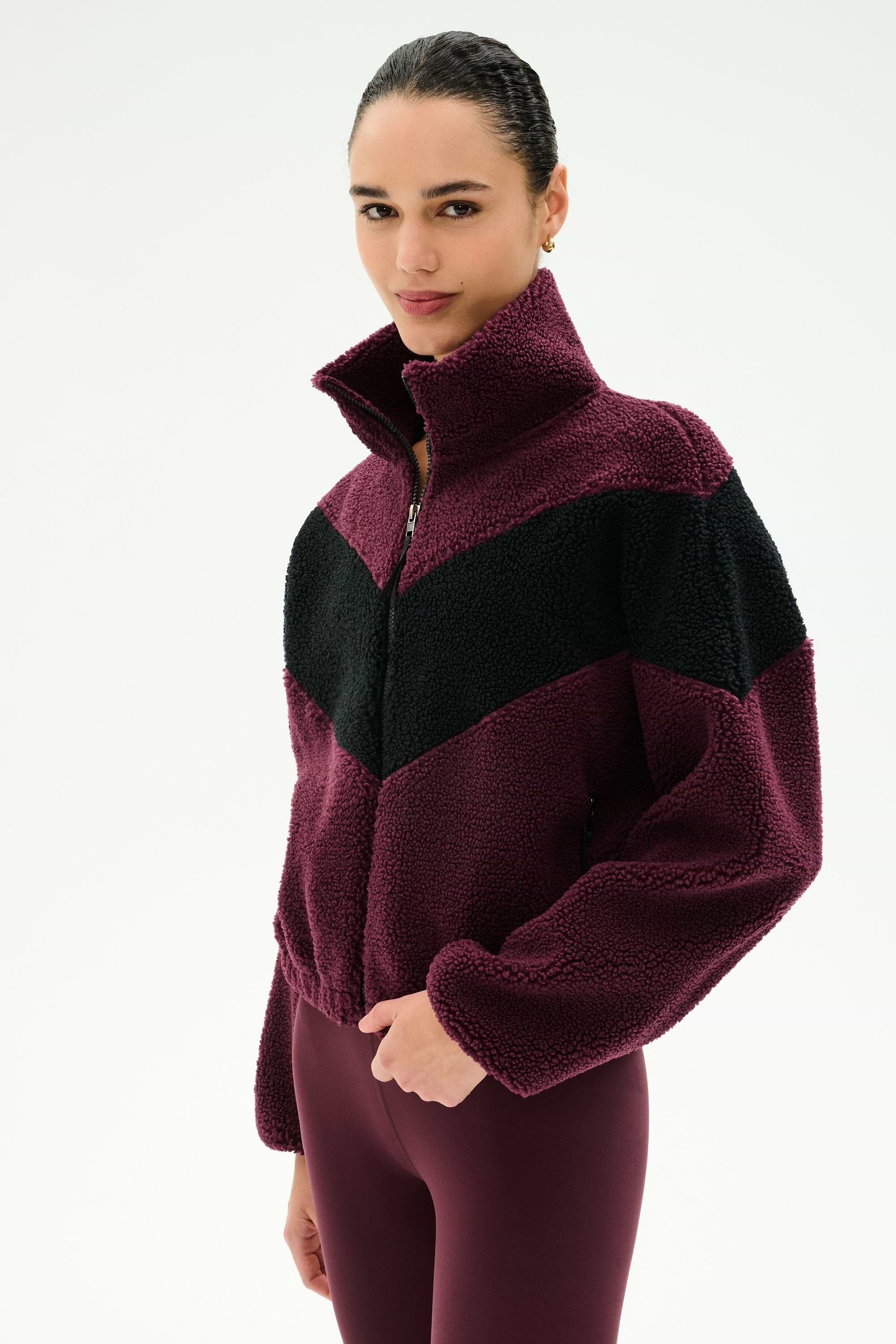 A person stands against a plain white background wearing the SPLITS59 Lottie Chevron Sherpa Full Zip Jacket in Dark Cherry/Black, crafted from soft 100% polyester fabric, paired with maroon leggings.