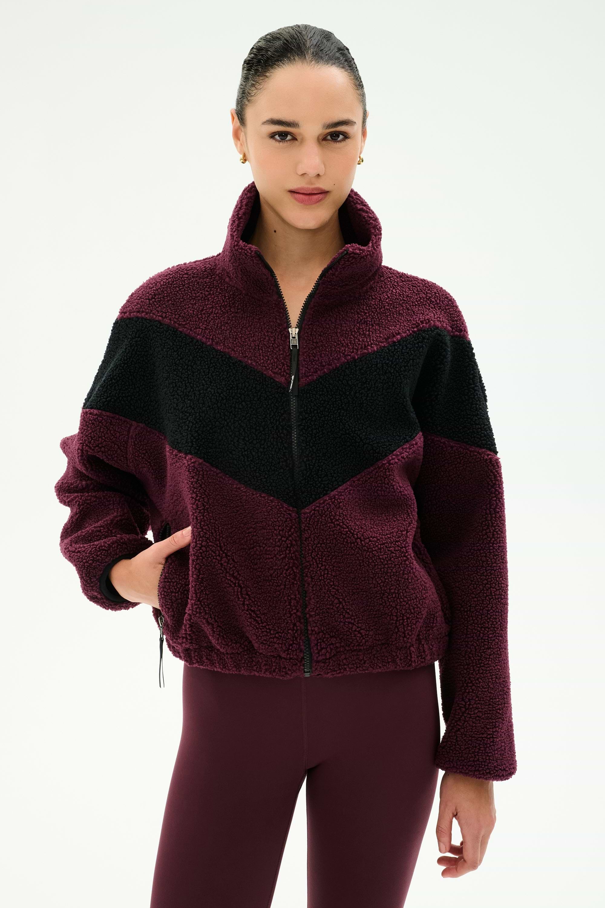 A woman stands against a plain light background wearing the SPLITS59 Lottie Chevron Sherpa Full Zip Jacket in Dark Cherry/Black, crafted from soft polyester, paired with matching maroon leggings.