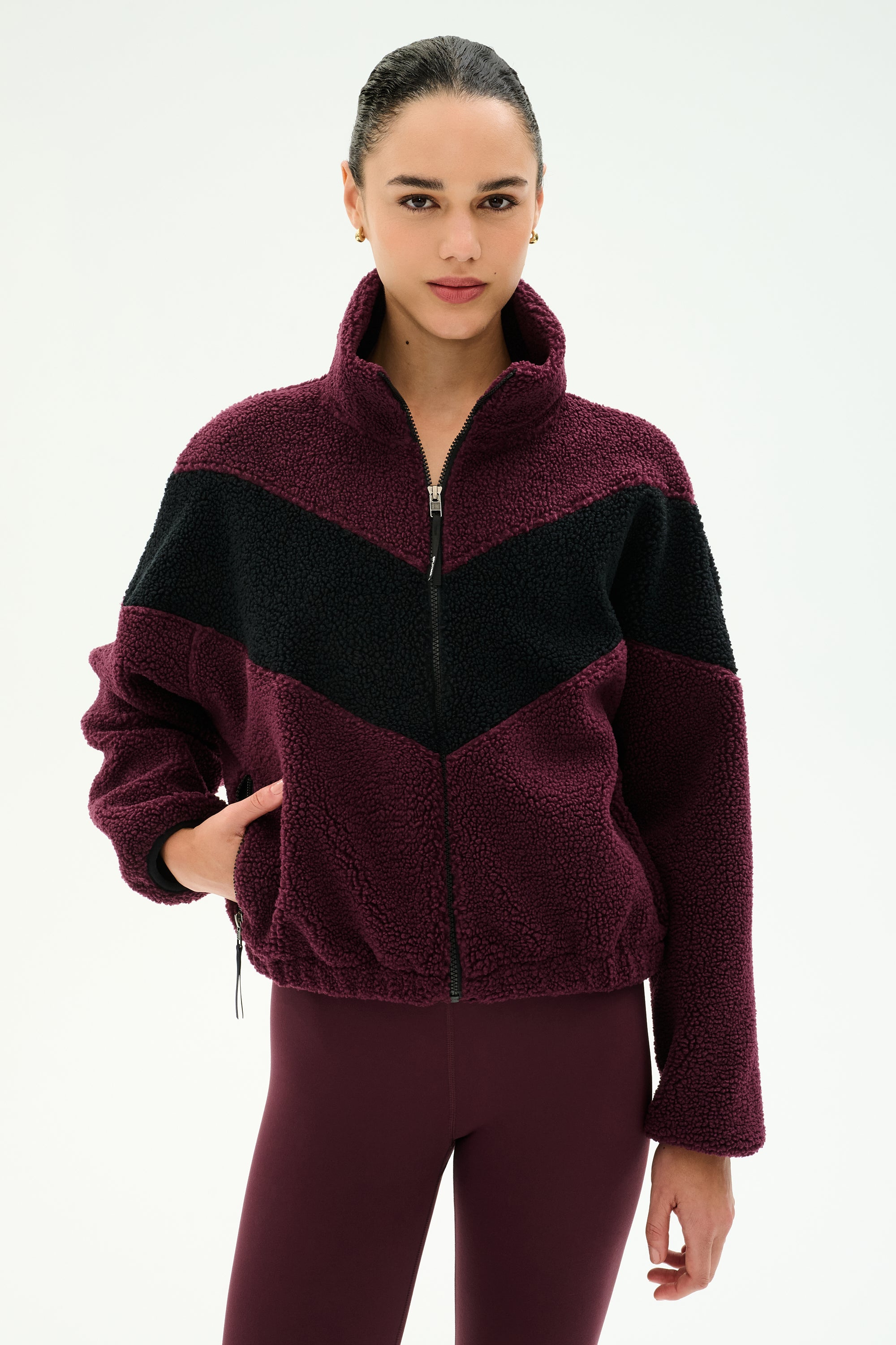 A woman stands against a plain light background wearing the SPLITS59 Lottie Chevron Sherpa Full Zip Jacket in Dark Cherry/Black, crafted from soft polyester, paired with matching maroon leggings.