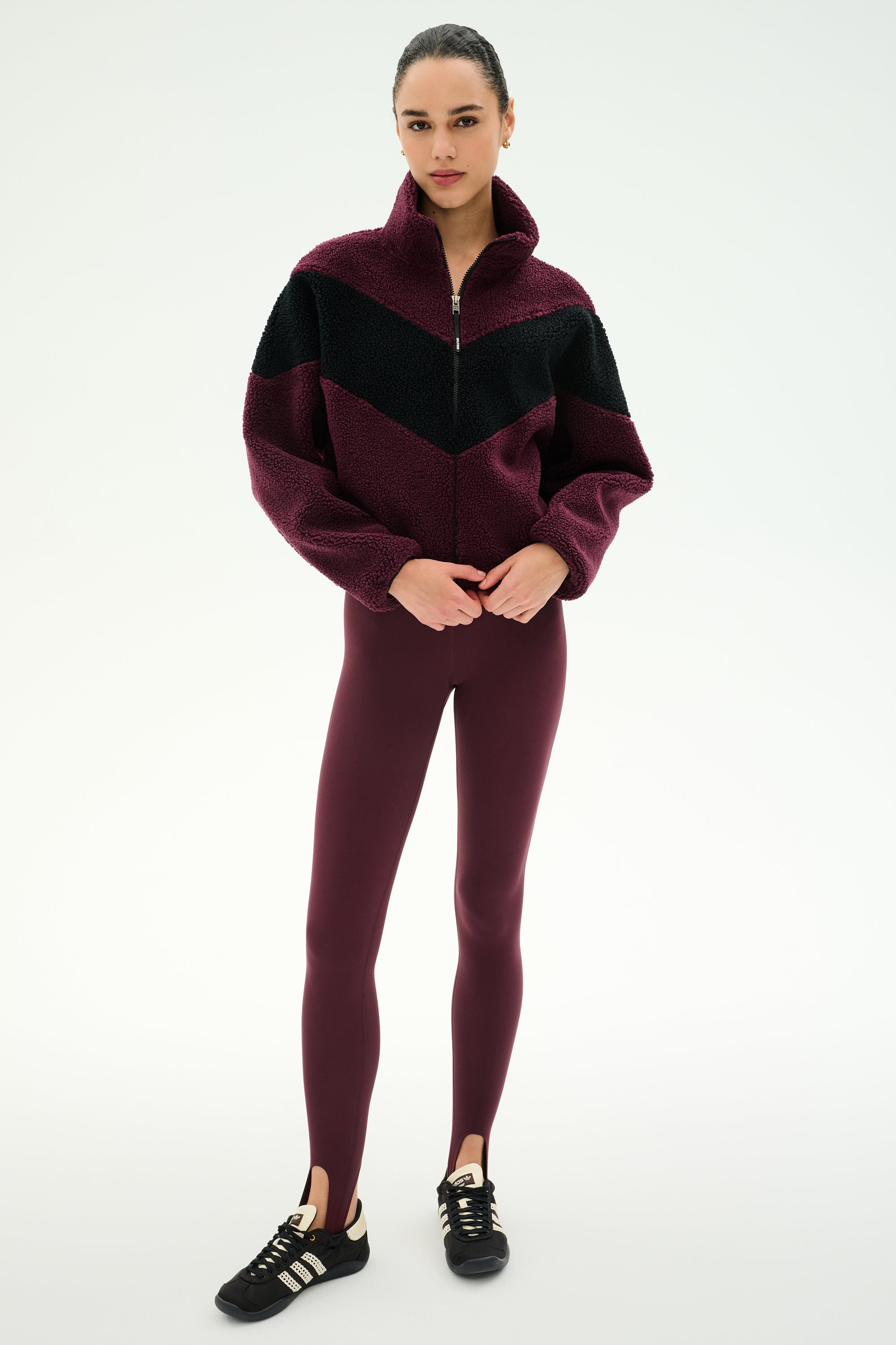 A woman stands facing forward against a plain white background, wearing the SPLITS59 Lottie Chevron Sherpa Full Zip Jacket in Dark Cherry/Black, paired with maroon stirrup leggings and black sneakers.