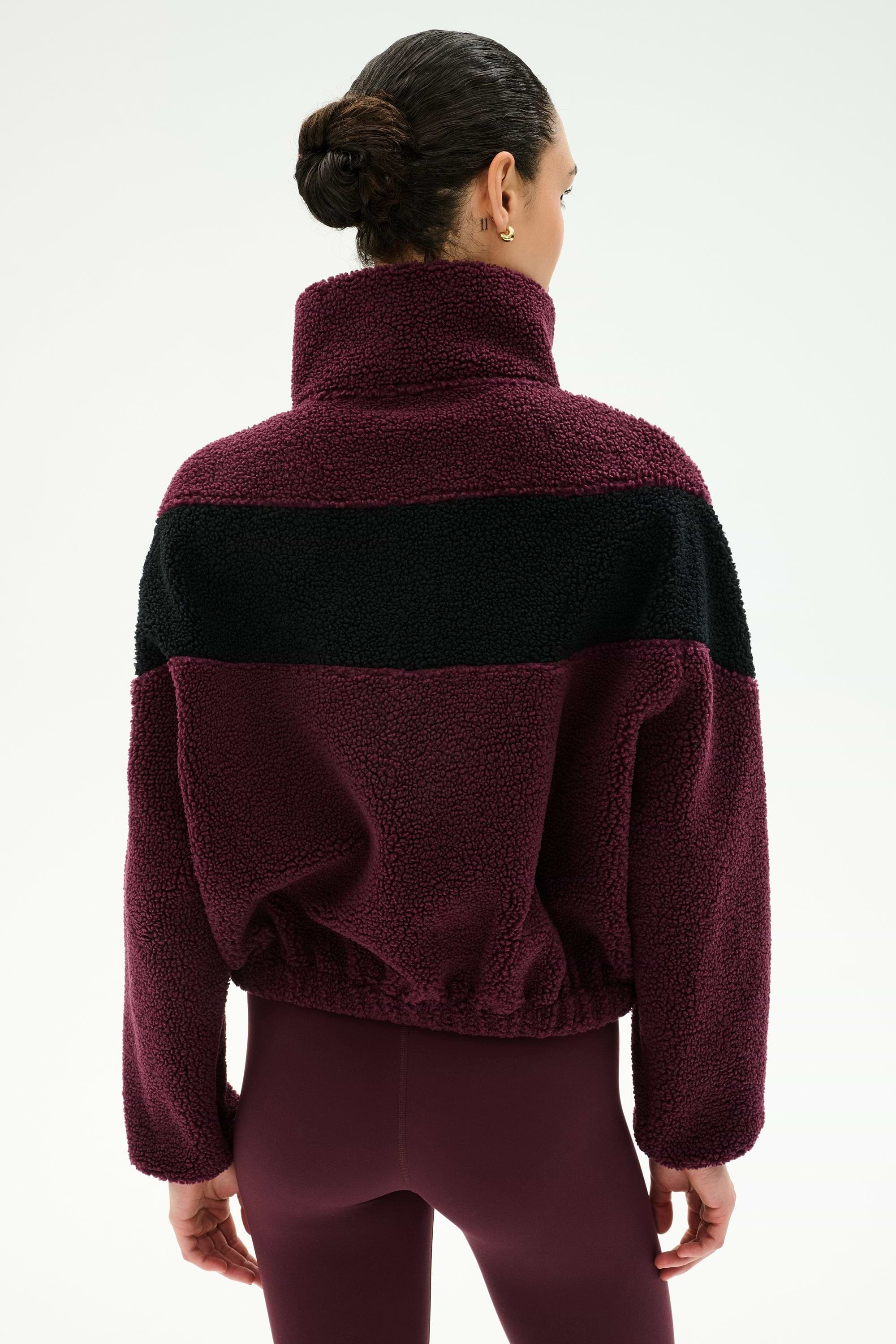 A person with dark hair in a bun is shown from the back against a plain background, wearing the SPLITS59 Lottie Chevron Sherpa Full Zip Jacket in Dark Cherry/Black—crafted from 100% polyester and featuring a black chevron stripe—paired with matching maroon leggings.