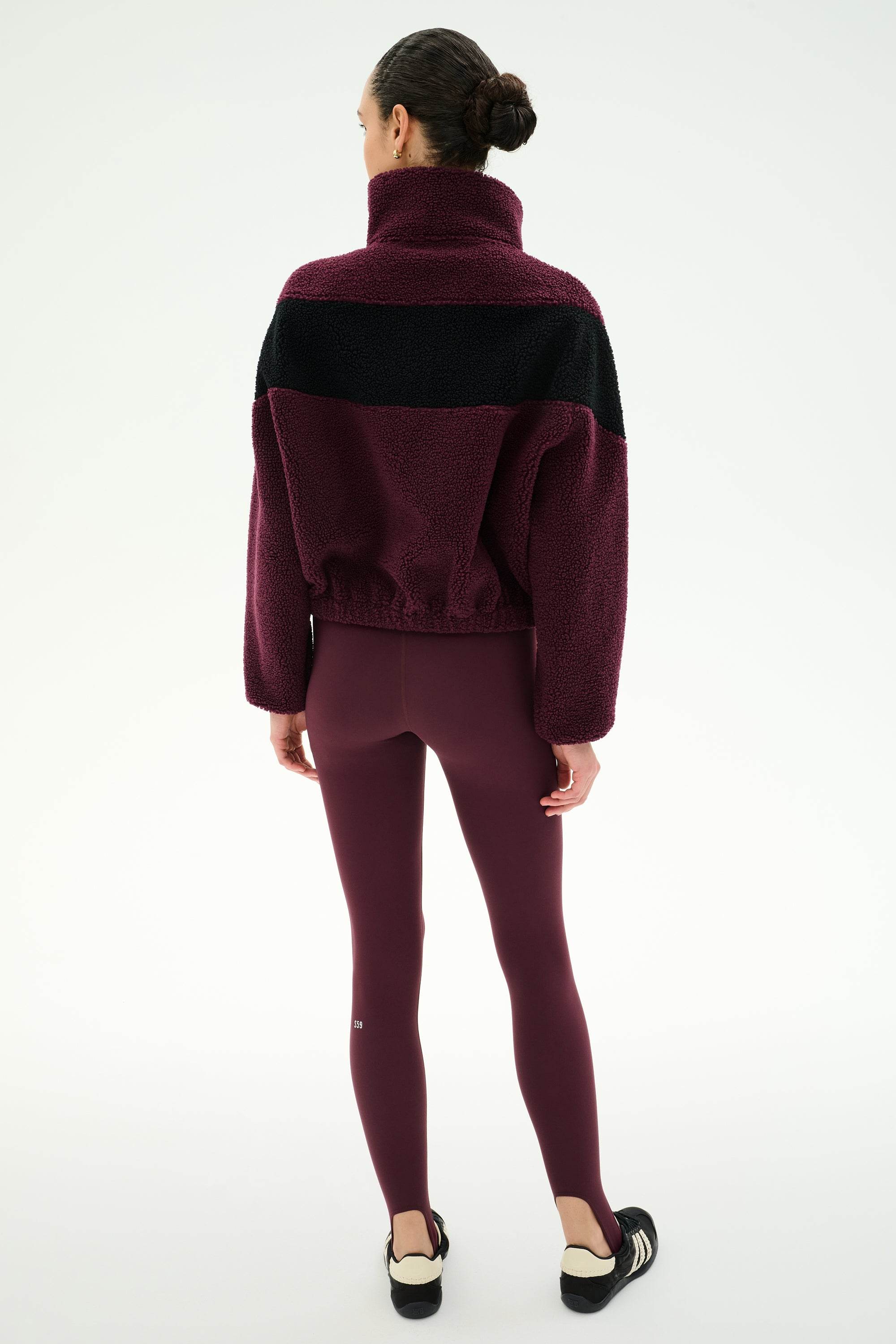 A woman stands facing away against a plain white background, wearing the SPLITS59 Lottie Chevron Sherpa Full Zip Jacket in Dark Cherry/Black—crafted from 100% polyester—with matching maroon leggings and black slip-on shoes.