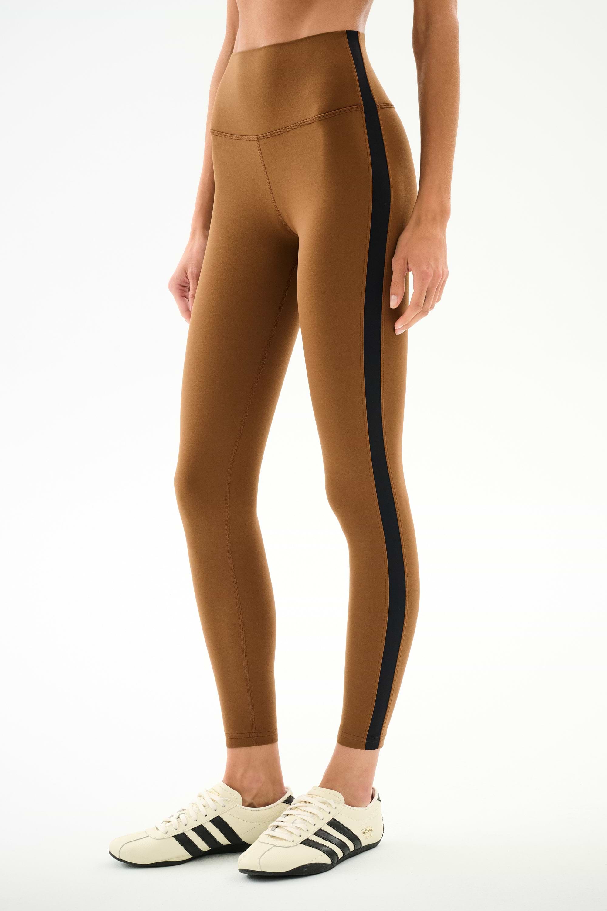 A person wears SPLITS59 Clare High Waist Rigor 7/8 - Toffee/Black leggings with cream sneakers featuring black stripes, standing against a white background.