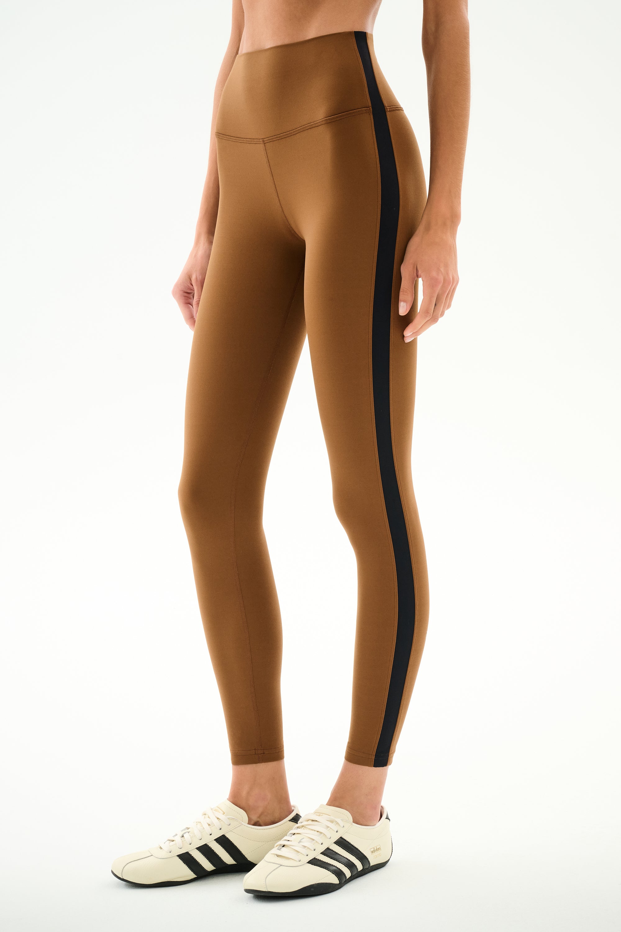 A person wears SPLITS59 Clare High Waist Rigor 7/8 - Toffee/Black leggings with cream sneakers featuring black stripes, standing against a white background.