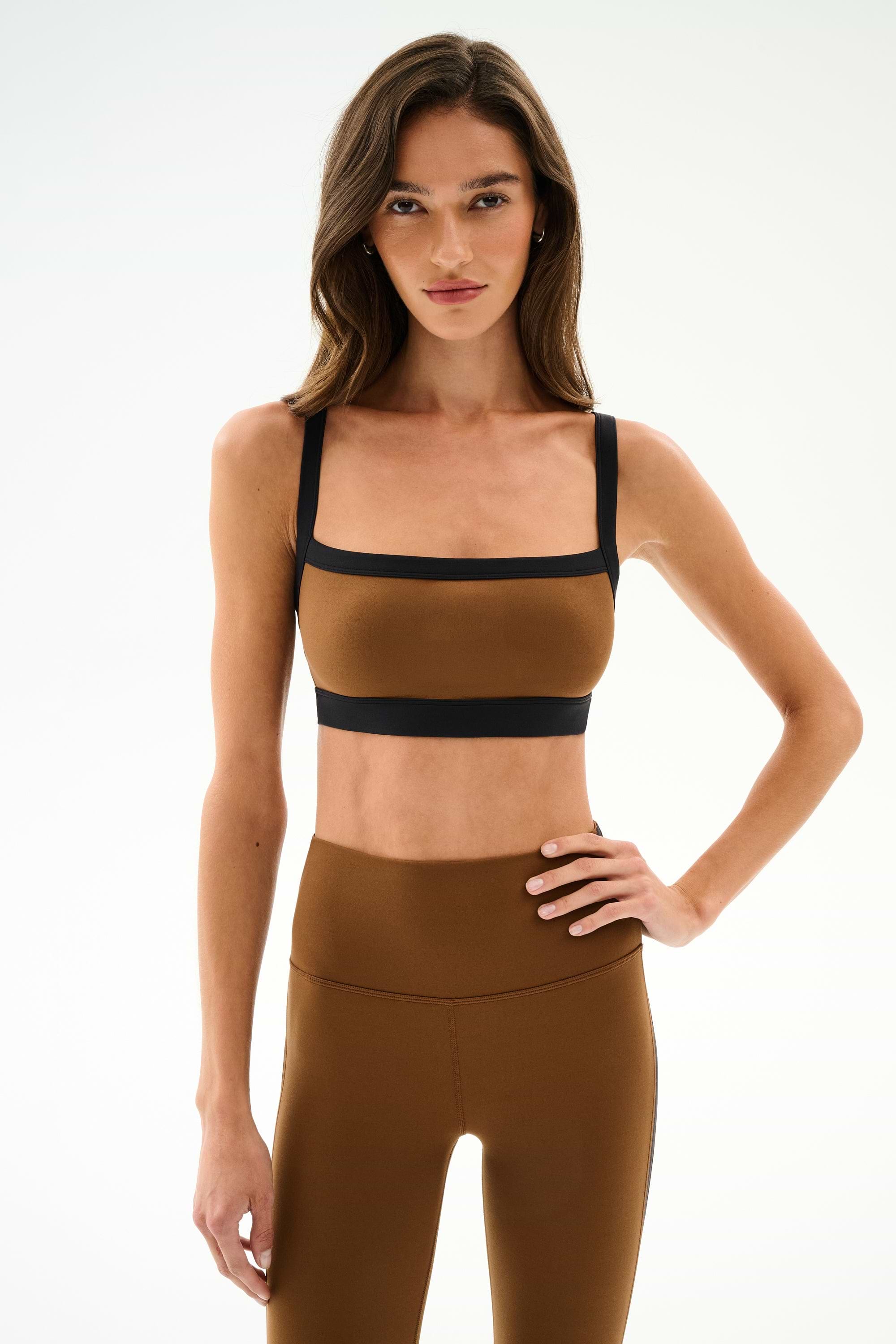 A woman wearing a Monah Rigor Bra -Toffee/Black sports bra with black trim and matching tan high-waisted leggings.