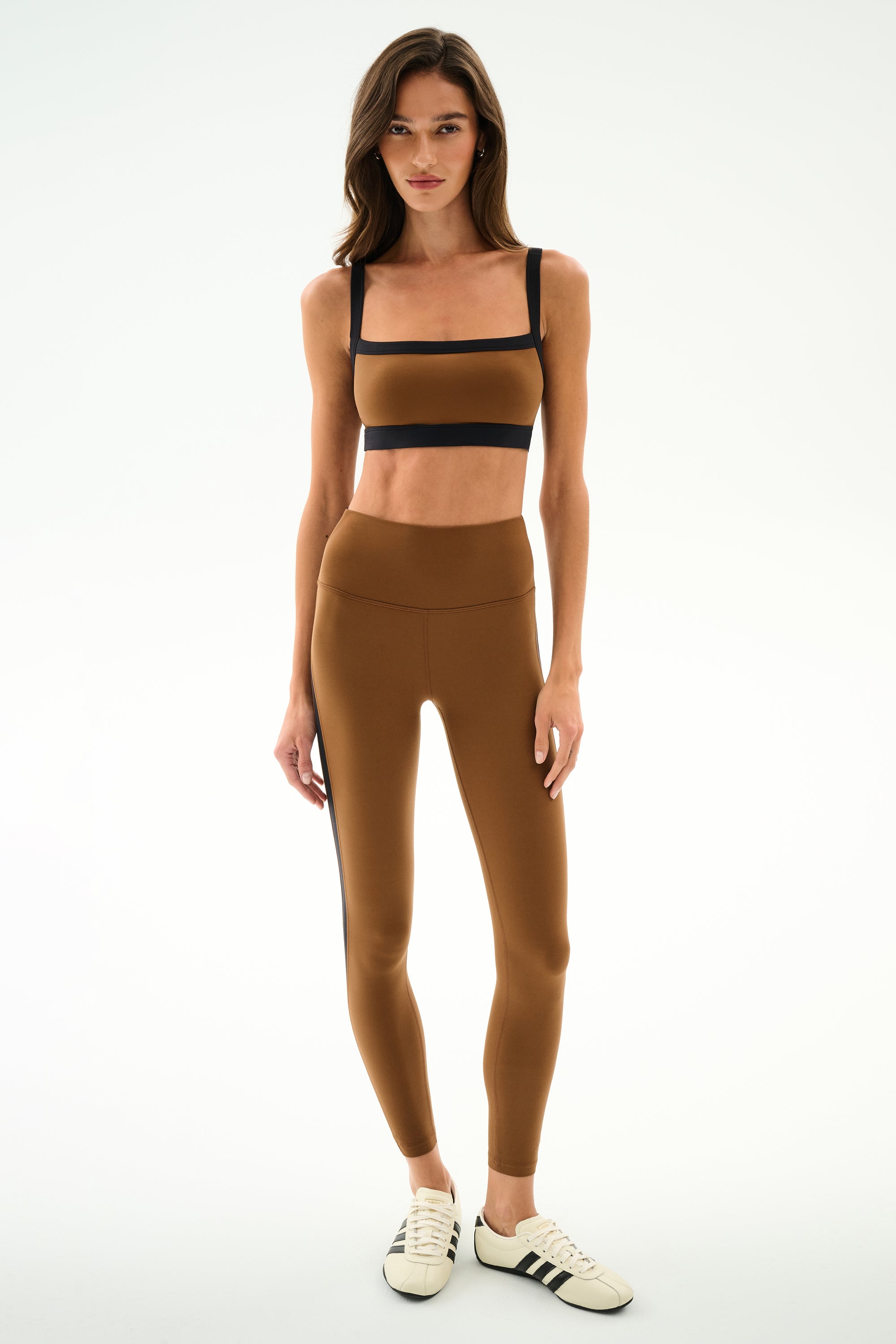 Woman faces forward in SPLITS59 Clare High Waist Rigor 7/8 - Toffee/Black leggings and sports bra, paired with white sneakers with black stripes, against a plain white background.