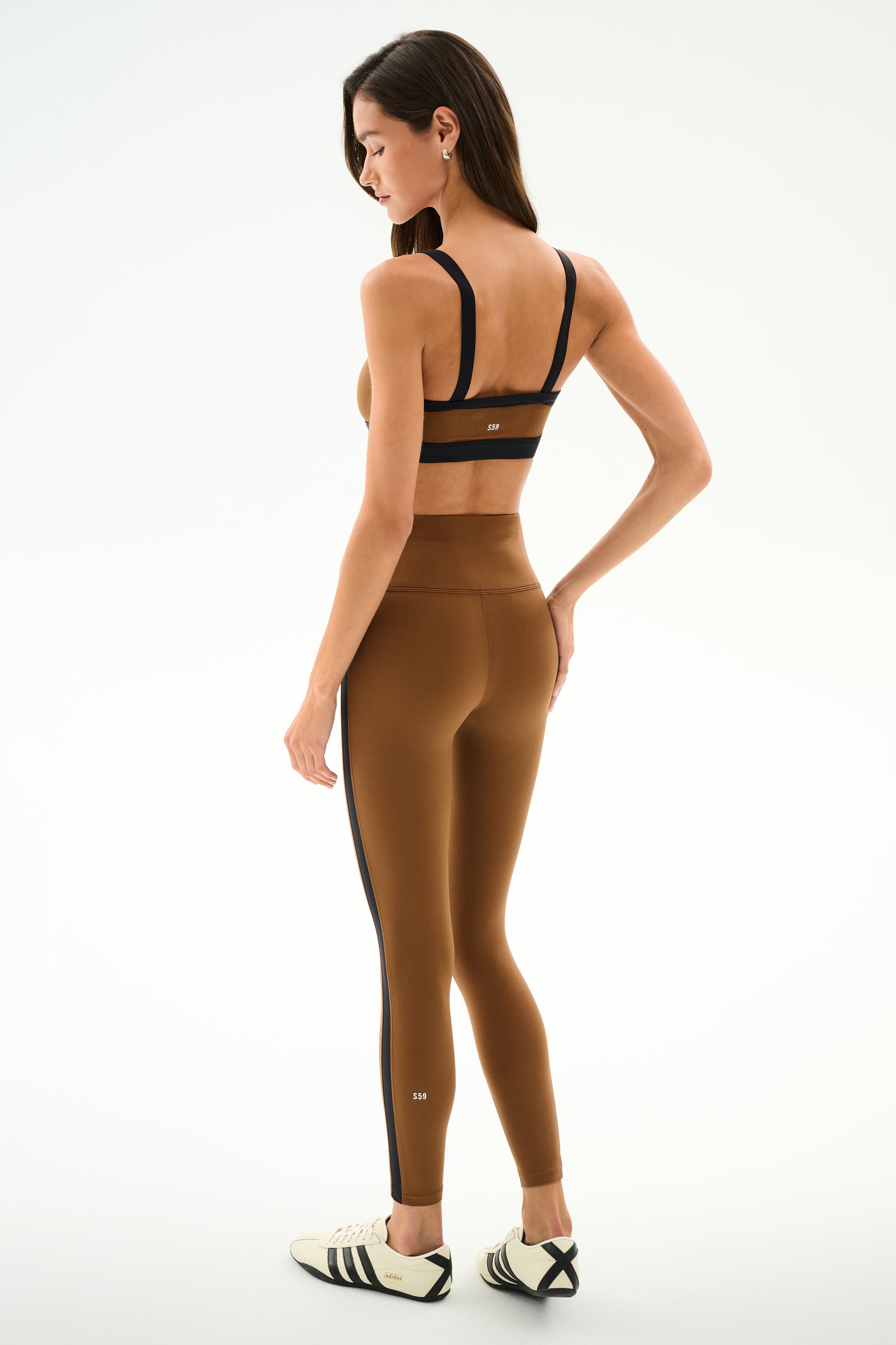 Woman wearing the SPLITS59 Monah Rigor Bra - Toffee/Black, featuring black stripes, and white sneakers with black stripes, standing sideways against a plain background.