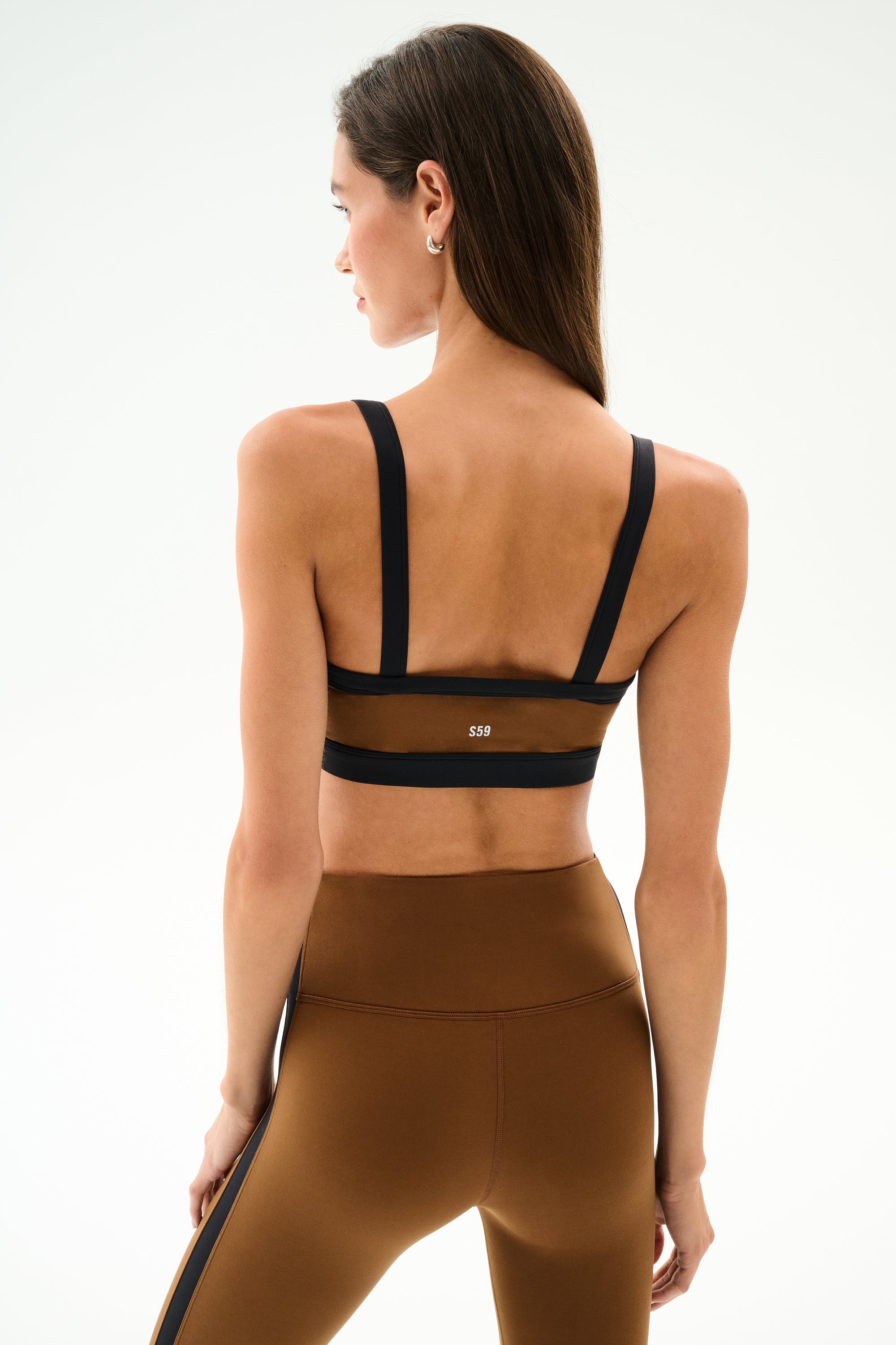 A woman faces away wearing the SPLITS59 Monah Rigor Bra - Toffee/Black with matching high-waisted leggings against a plain background.