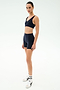 A woman poses against a plain white background wearing SPLITS59’s Airweight High Waist Short in Indigo with a matching workout top, white socks, and white athletic shoes.