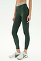 A person wears SPLITS59 Bailey Active Rib High Waist 7/8 leggings in Heather Military, featuring a striped waistband, paired with white athletic shoes, standing against a plain background.