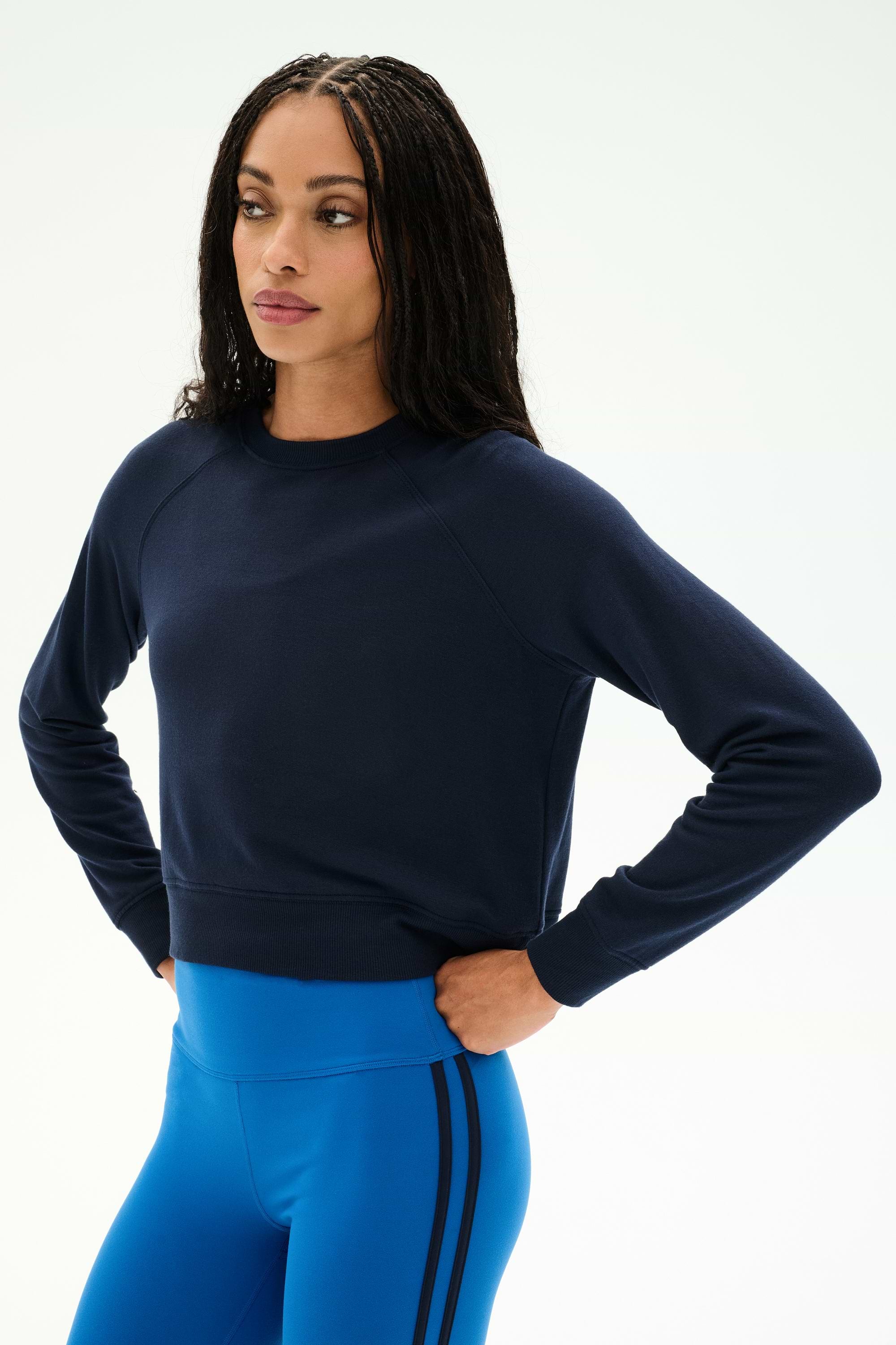 A woman stands with her hands on her hips against a plain light background, wearing the SPLITS59 Brooks Fleece Long Sleeve Sweatshirt in Indigo—a raglan sleeve crewneck pullover. She pairs it with bright blue athletic leggings featuring black stripes for a stylish look made in Los Angeles.