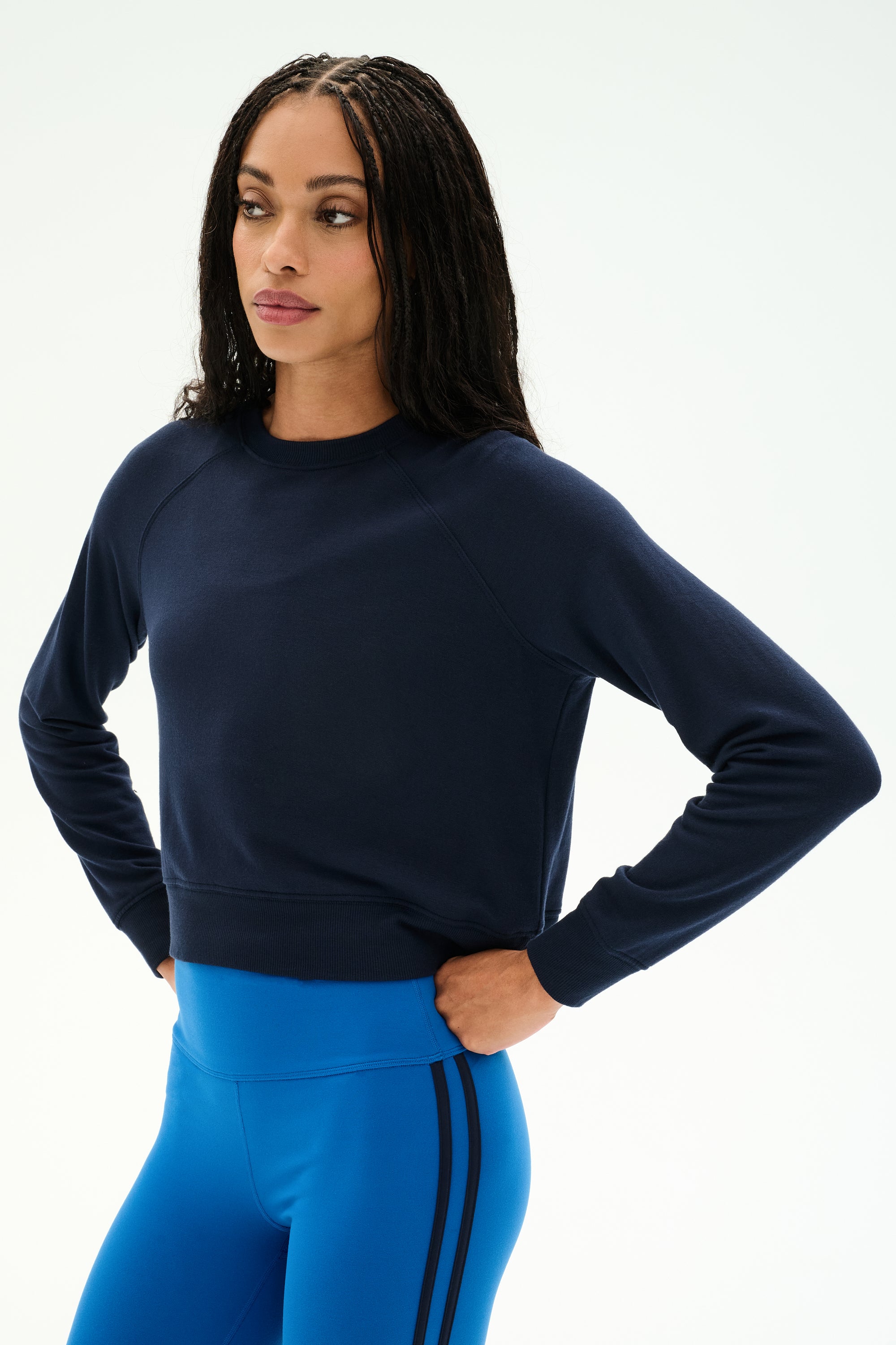 A woman stands with her hands on her hips against a plain light background, wearing the SPLITS59 Brooks Fleece Long Sleeve Sweatshirt in Indigo—a raglan sleeve crewneck pullover. She pairs it with bright blue athletic leggings featuring black stripes for a stylish look made in Los Angeles.