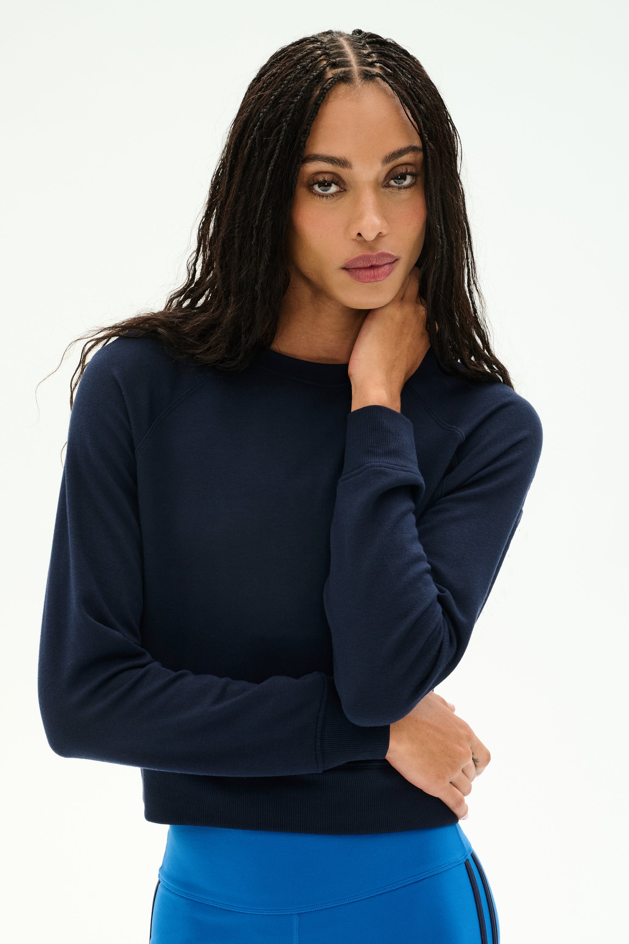 A woman with long braided hair is wearing the SPLITS59 Brooks Fleece Long Sleeve Sweatshirt in Indigo and blue leggings, standing against a plain white background with her hand touching her neck.