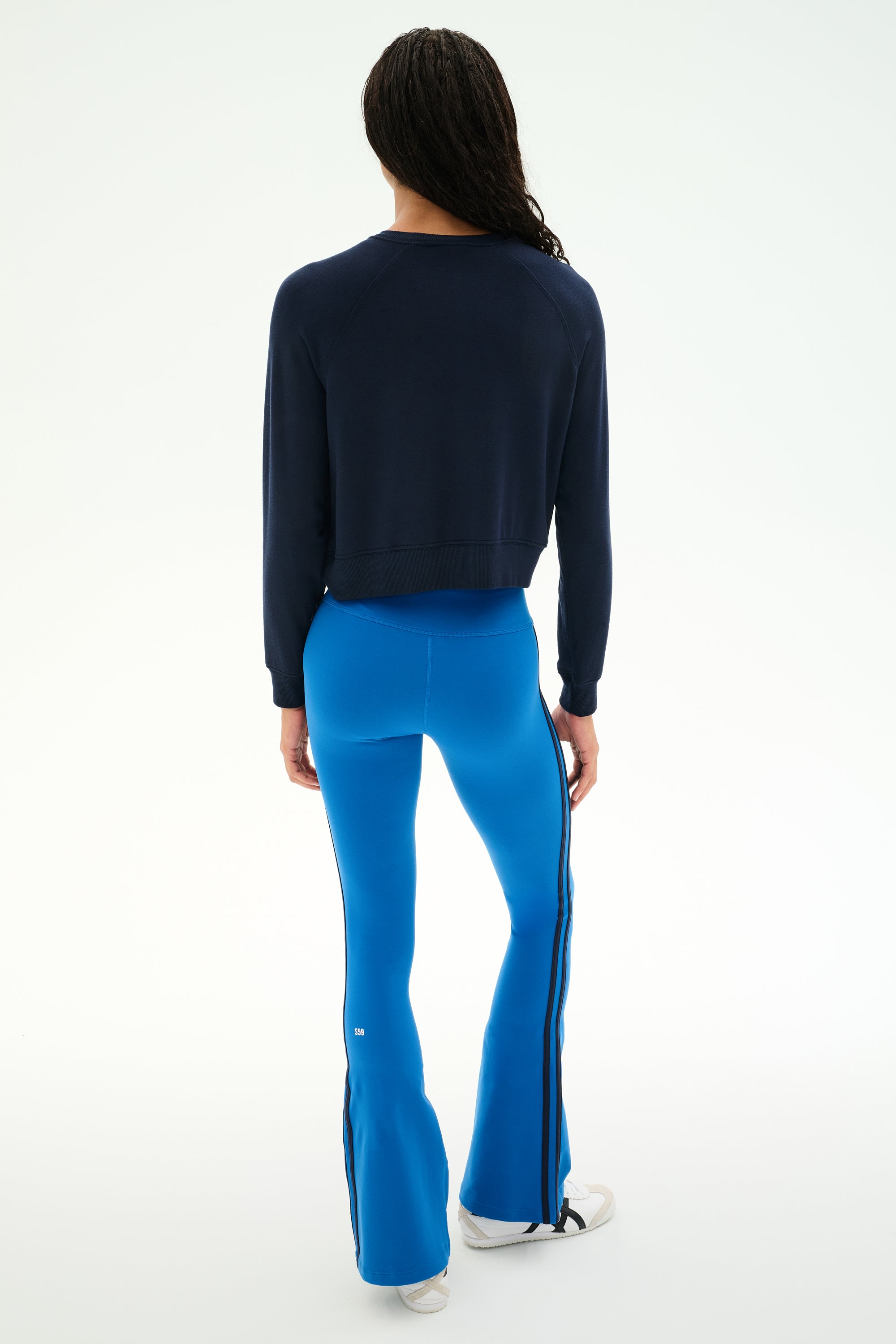 A person stands with their back to the camera, wearing the SPLITS59 Brooks Fleece Long Sleeve Sweatshirt in Indigo—a raglan sleeve crewneck pullover—paired with bright blue flared pants featuring black side stripes and white shoes, set against a plain light background. This stylish look is made in Los Angeles.