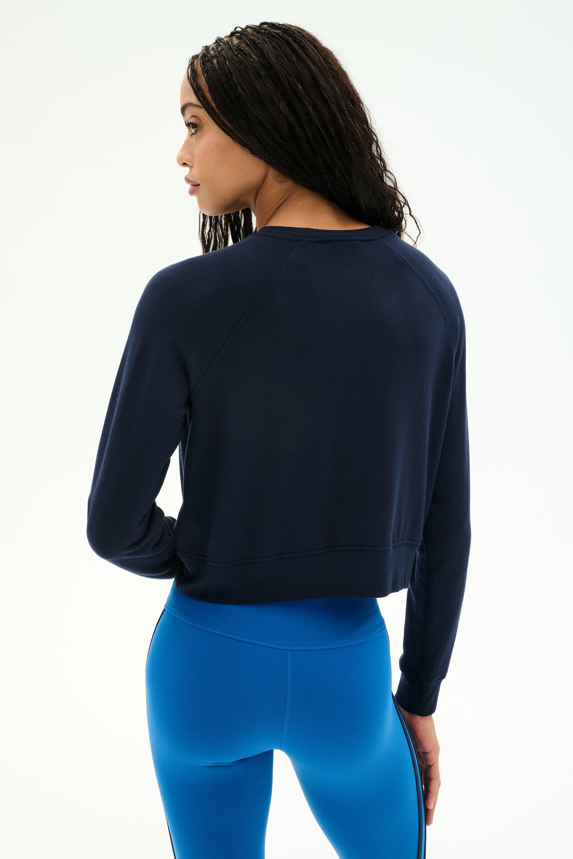 A person with long, braided hair is shown from the back against a plain white background, wearing the SPLITS59 Brooks Fleece Long Sleeve Sweatshirt in indigo and bright blue leggings. This stylish crewneck pullover features raglan sleeves and is made in Los Angeles.