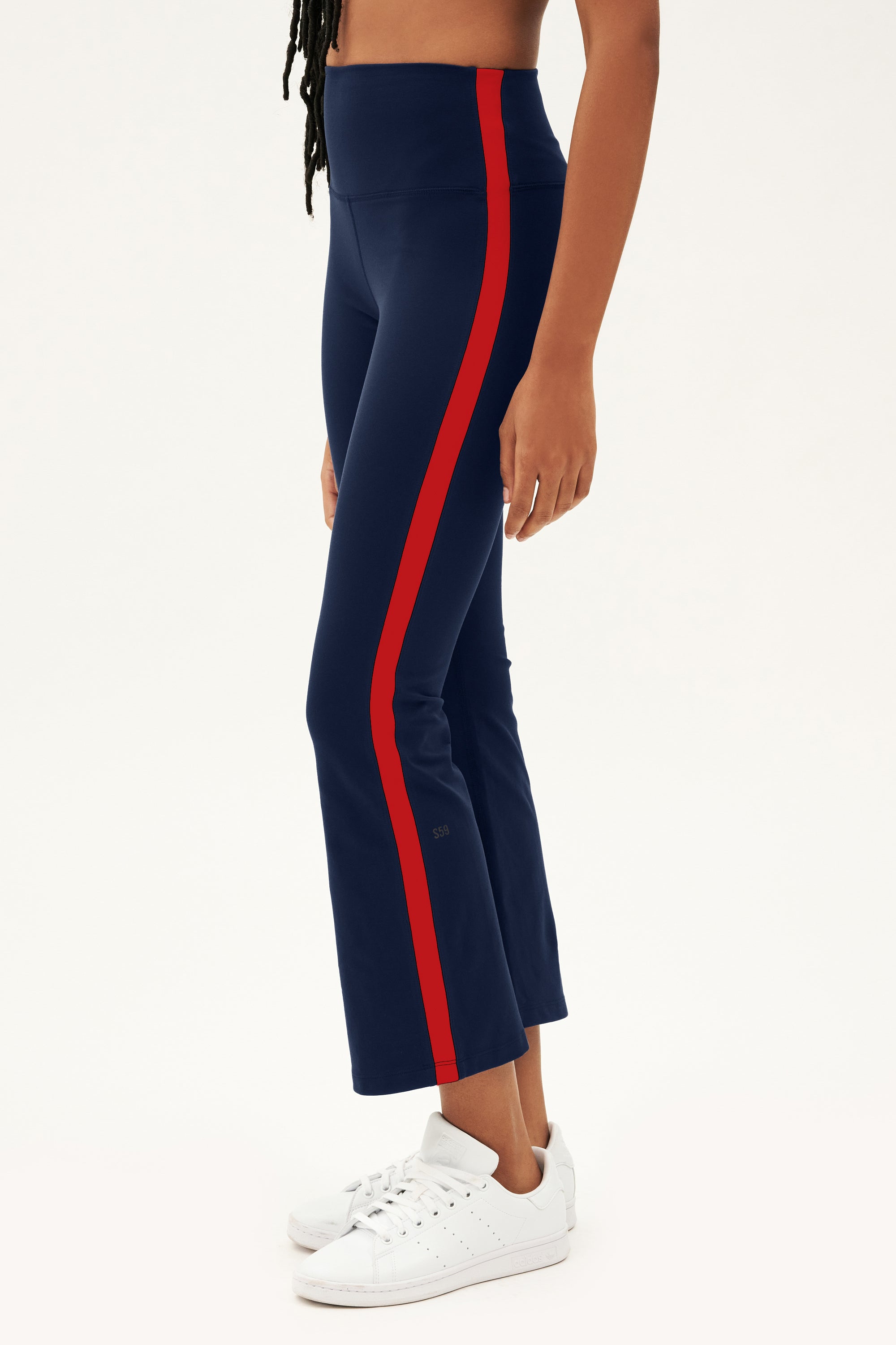 A person in profile wears SPLITS59 Raquel High Waist Crop - Indigo/Pirate Red, featuring a red side stripe and 4-way stretch supplex fabric, paired with white sneakers—ideal for yoga against a plain background.