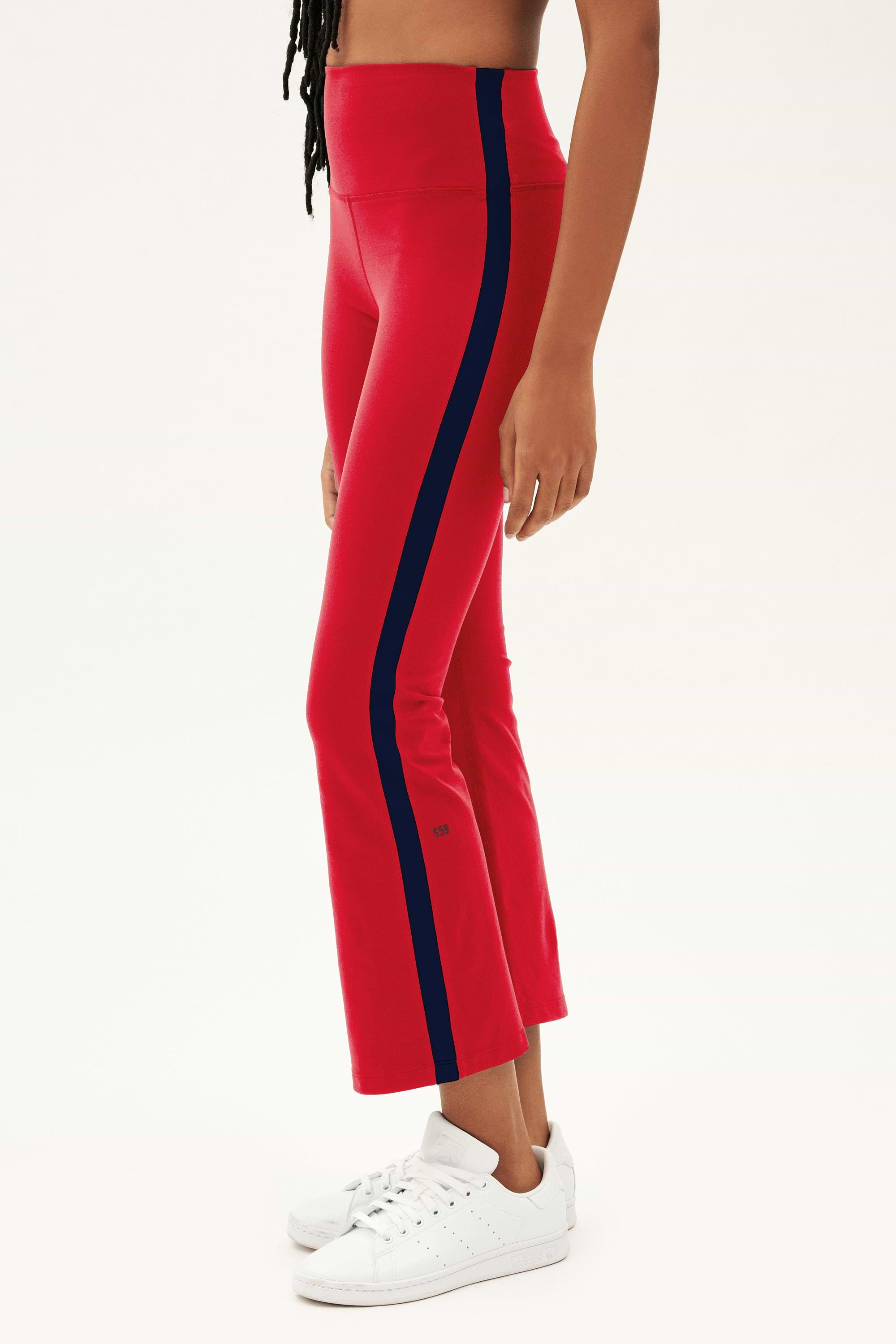 A person wears SPLITS59 Raquel High Waist Crop leggings in Pirate Red/Indigo, styled with white sneakers, standing before a plain white background.
