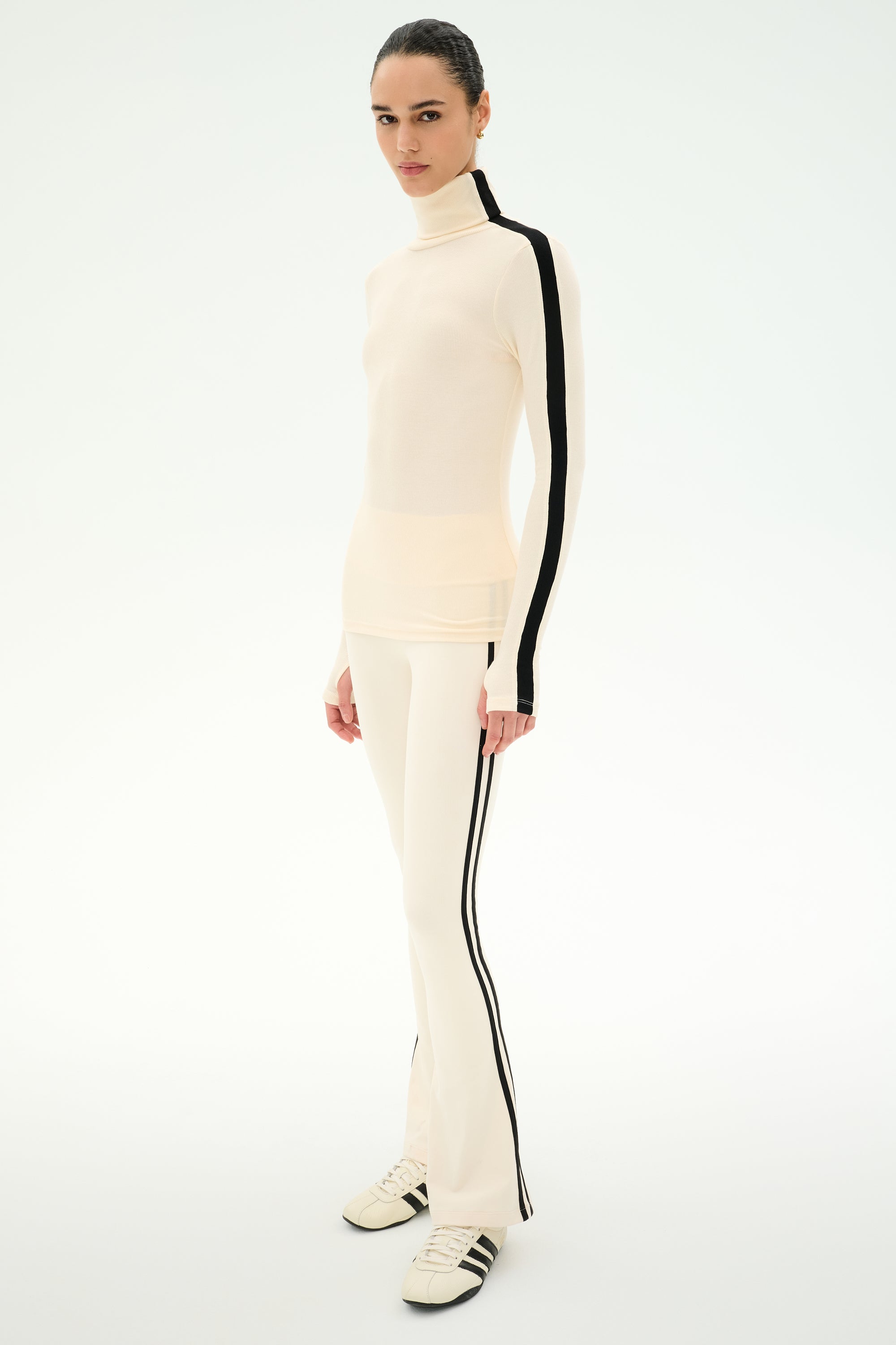Wearing the SPLITS59 Jackson Rib Full Length Turtleneck in Creme/Black, this person pairs the cream modal blend top featuring black side stripes with matching pants and cream sneakers, standing against a light background.