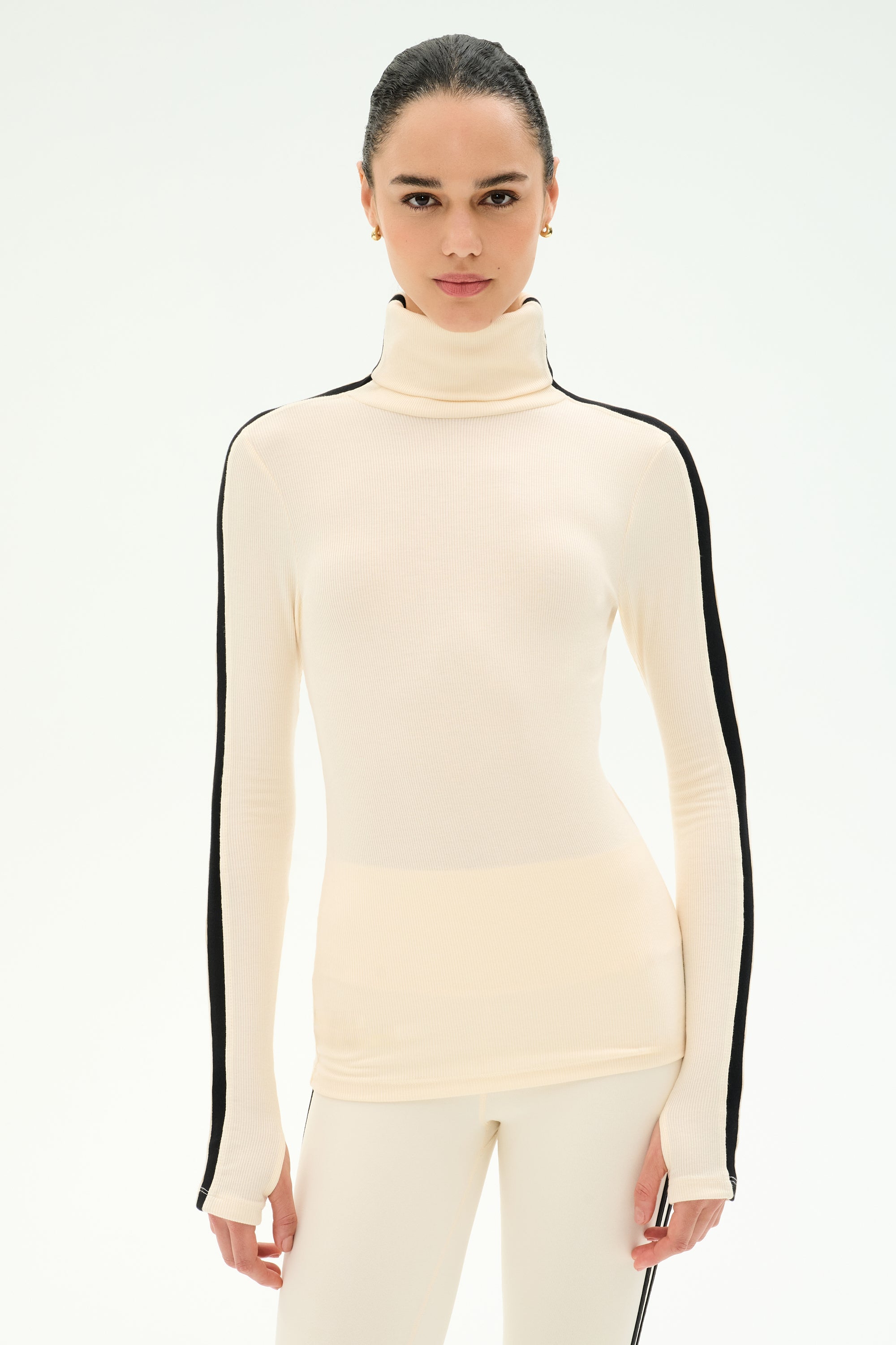 A woman models the SPLITS59 Jackson Rib Full Length Turtleneck in Creme/Black, featuring black stripes down the sleeves and crafted from a soft modal blend for ultimate comfort, standing against a plain white background.