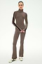 A woman stands against a plain light background wearing the SPLITS59 Raquel High Waist Supplex Flare pants in Lentil/Black, paired with a matching taupe turtleneck top and white sneakers. The soft nylon-spandex blend outfit features striking black side stripes.