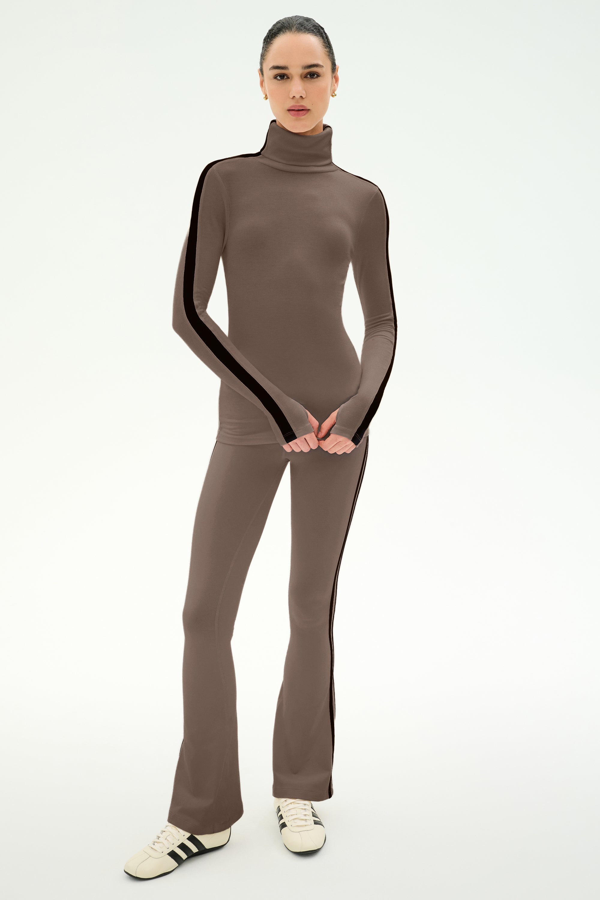 A woman stands against a plain light background wearing the SPLITS59 Raquel High Waist Supplex Flare pants in Lentil/Black, paired with a matching taupe turtleneck top and white sneakers. The soft nylon-spandex blend outfit features striking black side stripes.