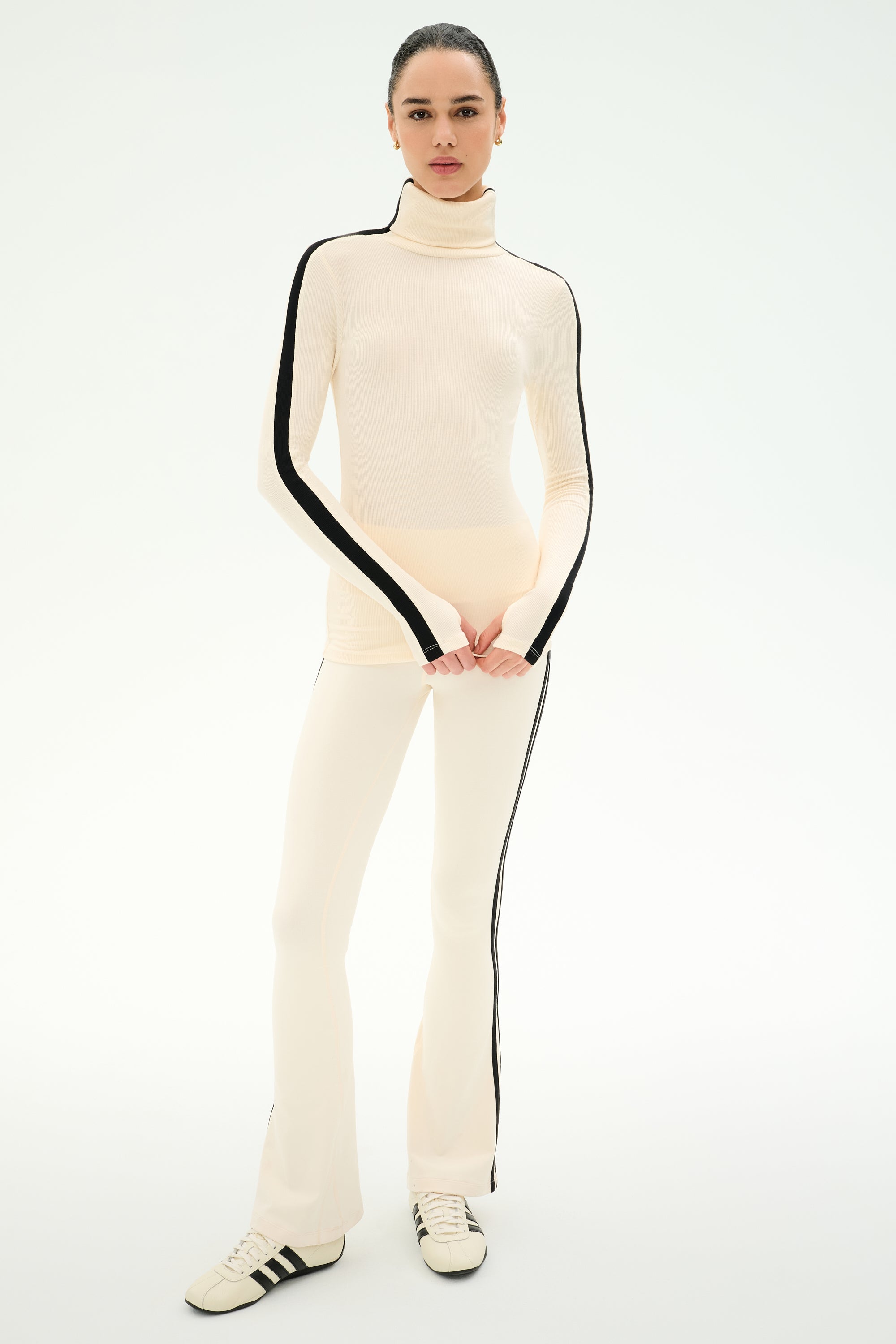 A woman stands on a plain background wearing the SPLITS59 Raquel High Waist Supplex Flare in Creme/Black—a cream turtleneck top paired with matching high waist flare leggings featuring black stripes down the sides and 4-way stretch for maximum comfort—styled with white sneakers.