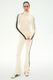 A woman stands on a plain background wearing the SPLITS59 Raquel High Waist Supplex Flare in Creme/Black—a cream turtleneck top paired with matching high waist flare leggings featuring black stripes down the sides and 4-way stretch for maximum comfort—styled with white sneakers.