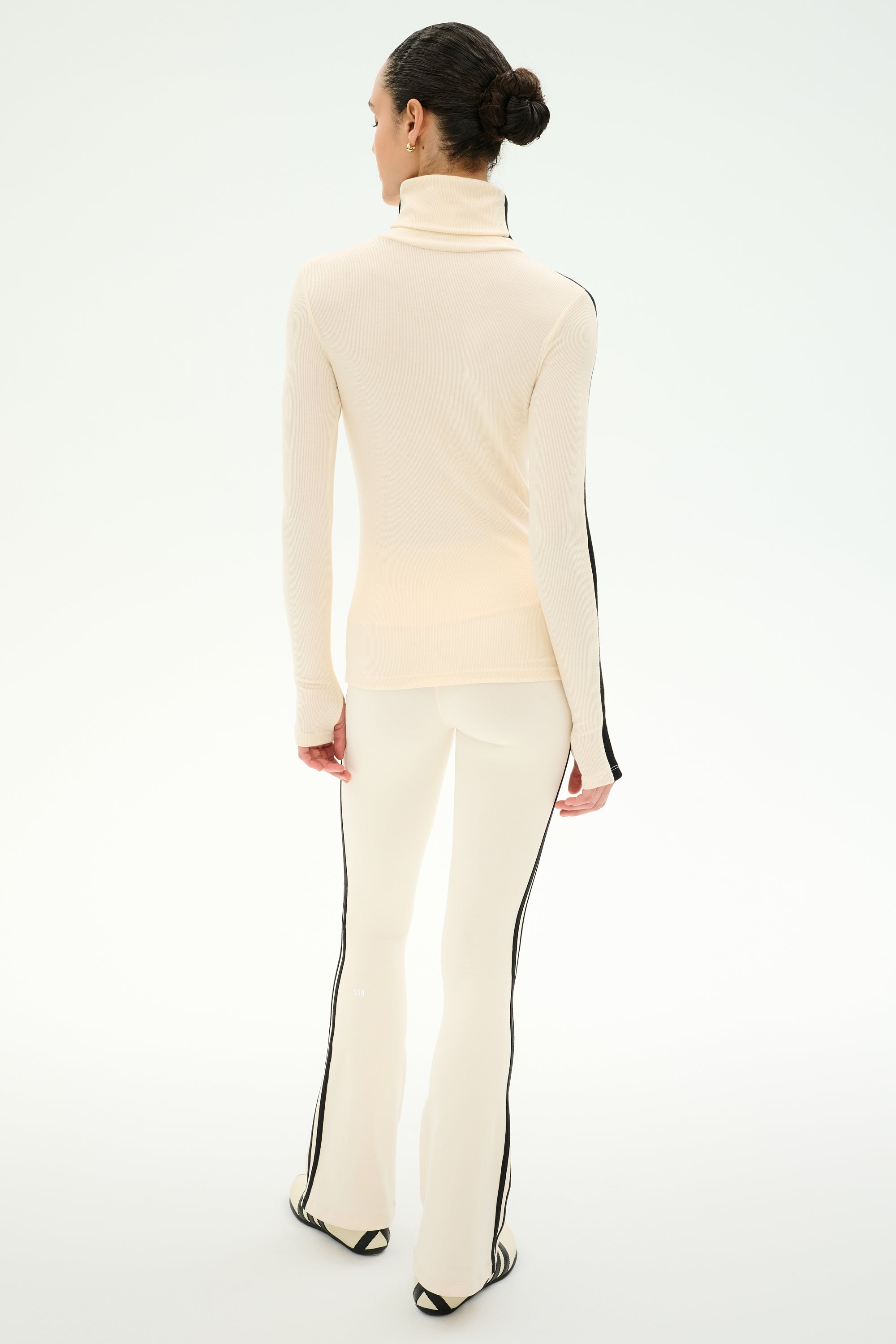A person stands facing away against a plain light background, wearing the SPLITS59 Jackson Rib Full Length Turtleneck in Creme/Black—a soft modal blend top featuring black side stripes.