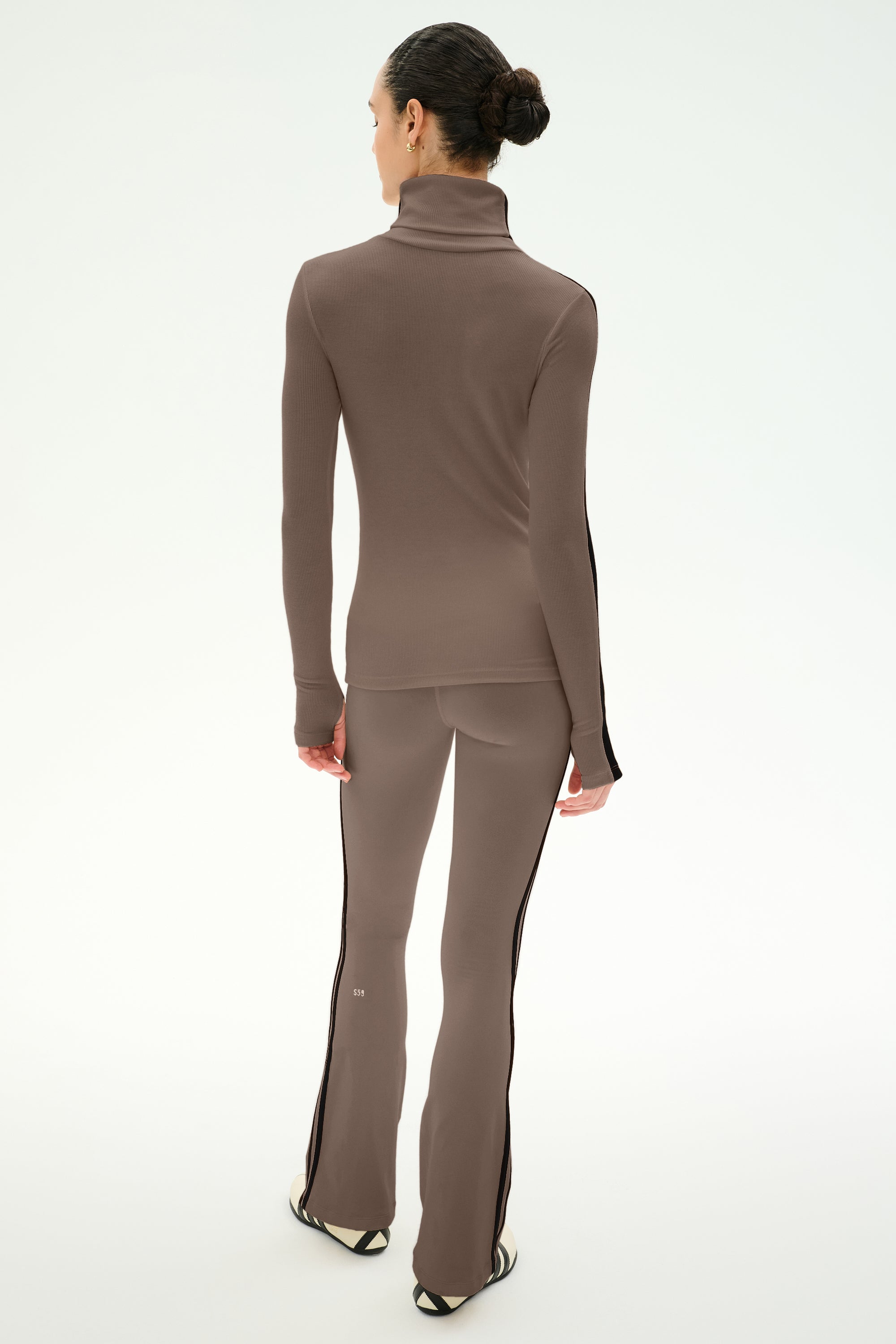 A person stands facing away, wearing the SPLITS59 Raquel High Waist Supplex Flare pants in Lentil/Black, styled with a fitted taupe turtleneck, against a plain white background.