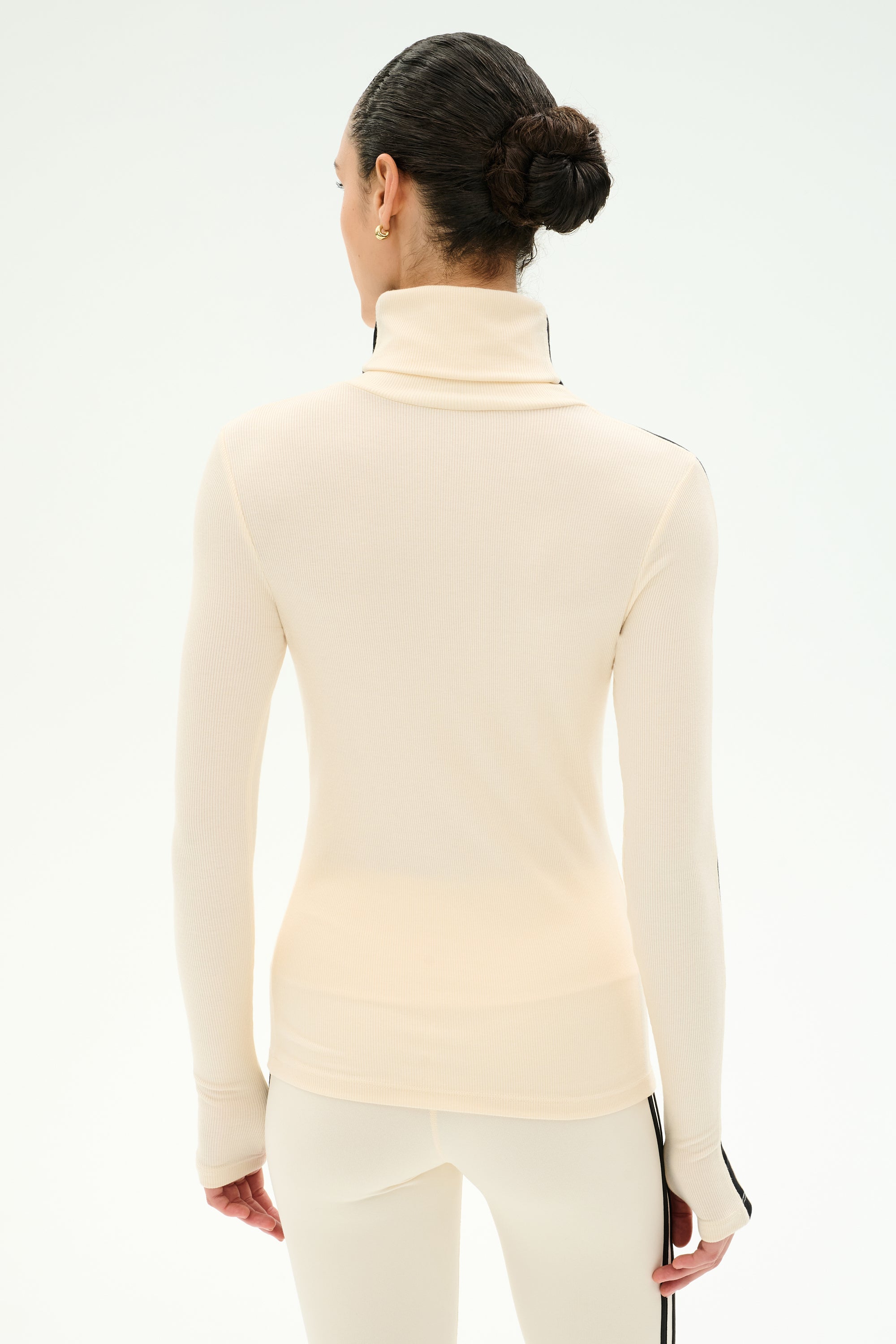 A person with dark hair styled in a bun is shown from the back against a plain white background, wearing the SPLITS59 Jackson Rib Full Length Turtleneck in Creme/Black, paired with matching pants; both pieces are crafted from soft modal blend fabric and feature long sleeves and a contemporary fit.