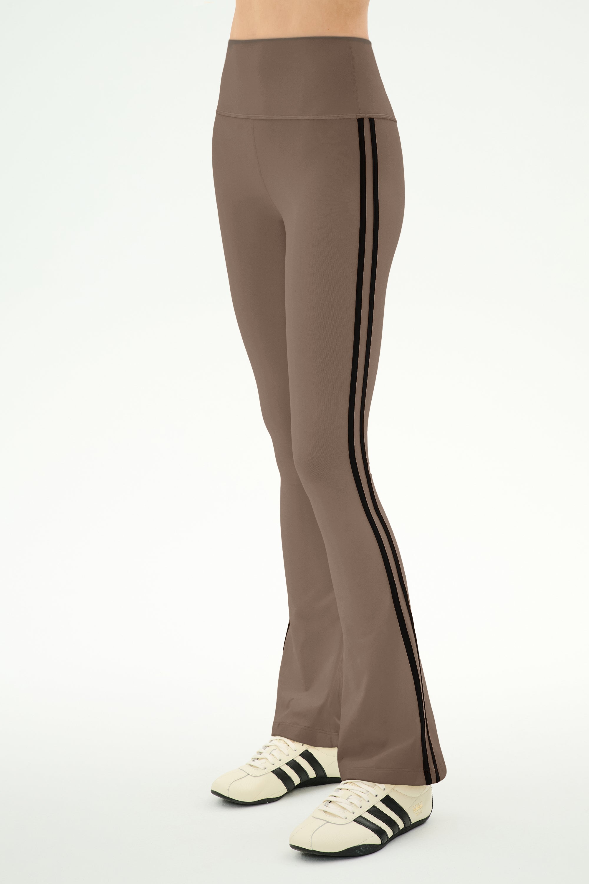 A person wearing the SPLITS59 Raquel High Waist Supplex Flare pants in Lentil/Black—a comfortable high-waisted brown flare style crafted from a nylon-spandex blend and detailed with black side stripes—paired with white sneakers featuring black stripes, stands against a plain light background.