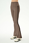 A person wearing the SPLITS59 Raquel High Waist Supplex Flare pants in Lentil/Black—a comfortable high-waisted brown flare style crafted from a nylon-spandex blend and detailed with black side stripes—paired with white sneakers featuring black stripes, stands against a plain light background.
