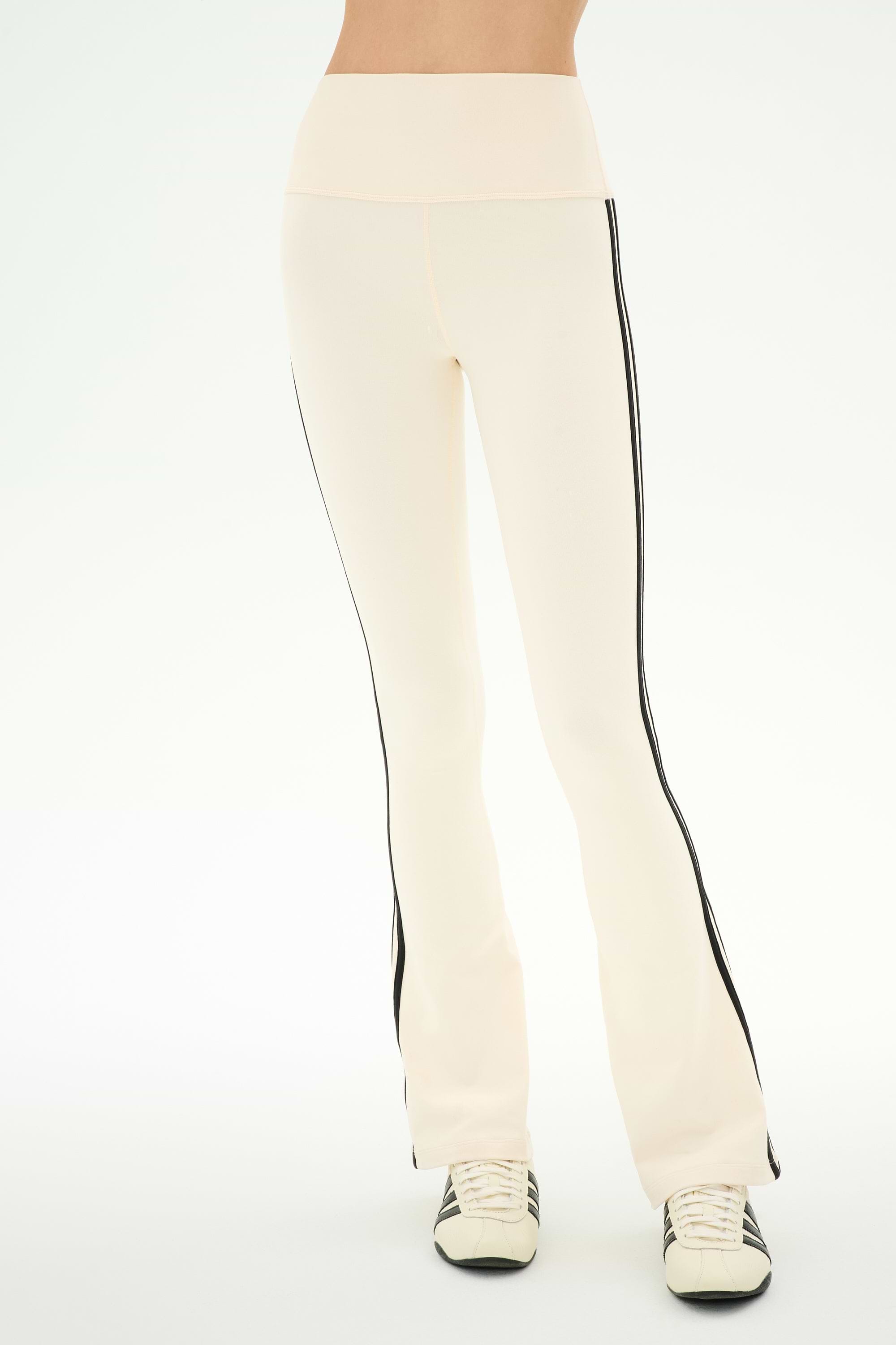 Wearing the SPLITS59 Raquel High Waist Supplex Flare pants in Creme/Black—featuring a cream color, high waist, black side stripes, and 4-way stretch Supplex fabric ideal for workouts—and white sneakers with black accents, this person stands against a plain light background.