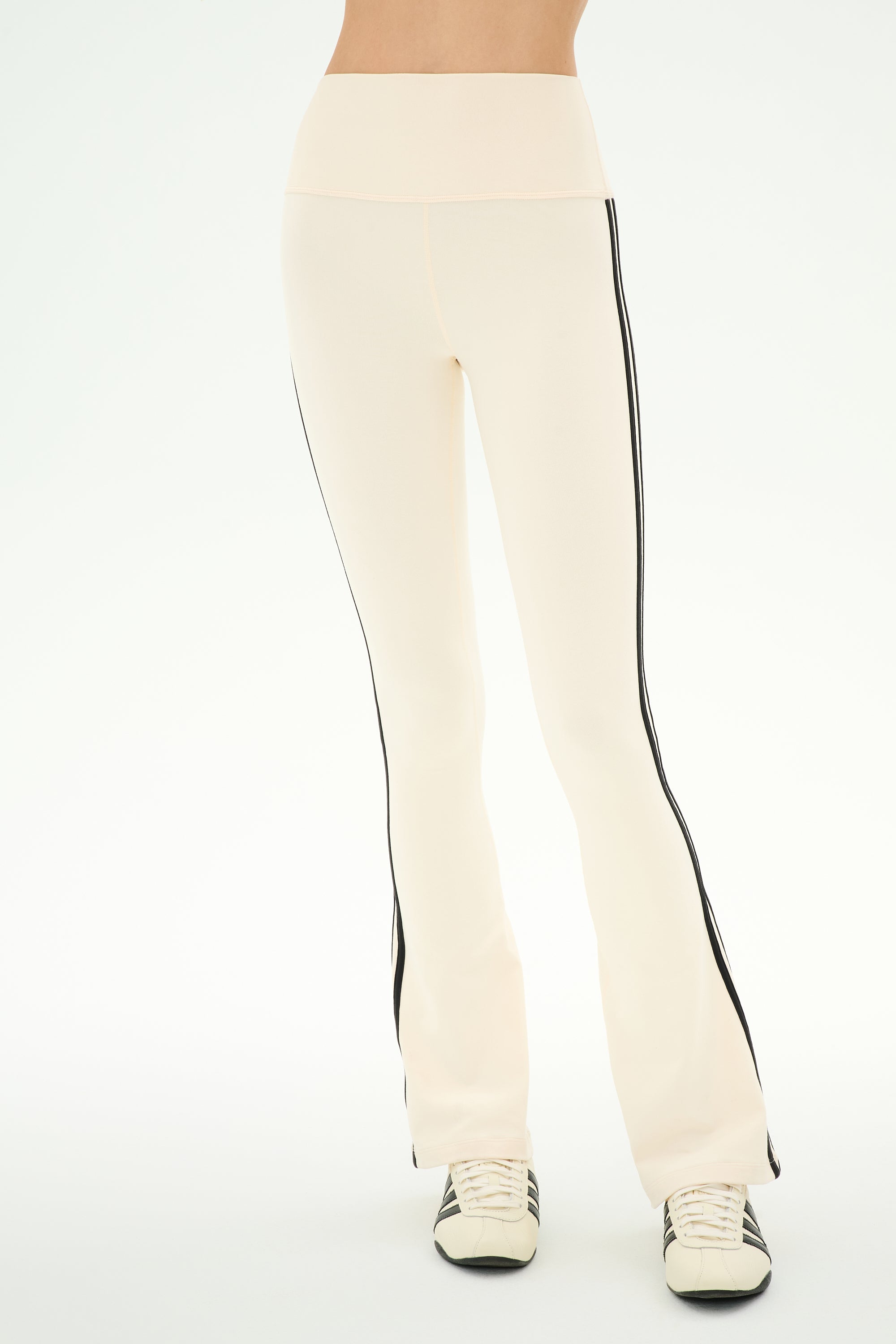 Wearing the SPLITS59 Raquel High Waist Supplex Flare pants in Creme/Black—featuring a cream color, high waist, black side stripes, and 4-way stretch Supplex fabric ideal for workouts—and white sneakers with black accents, this person stands against a plain light background.