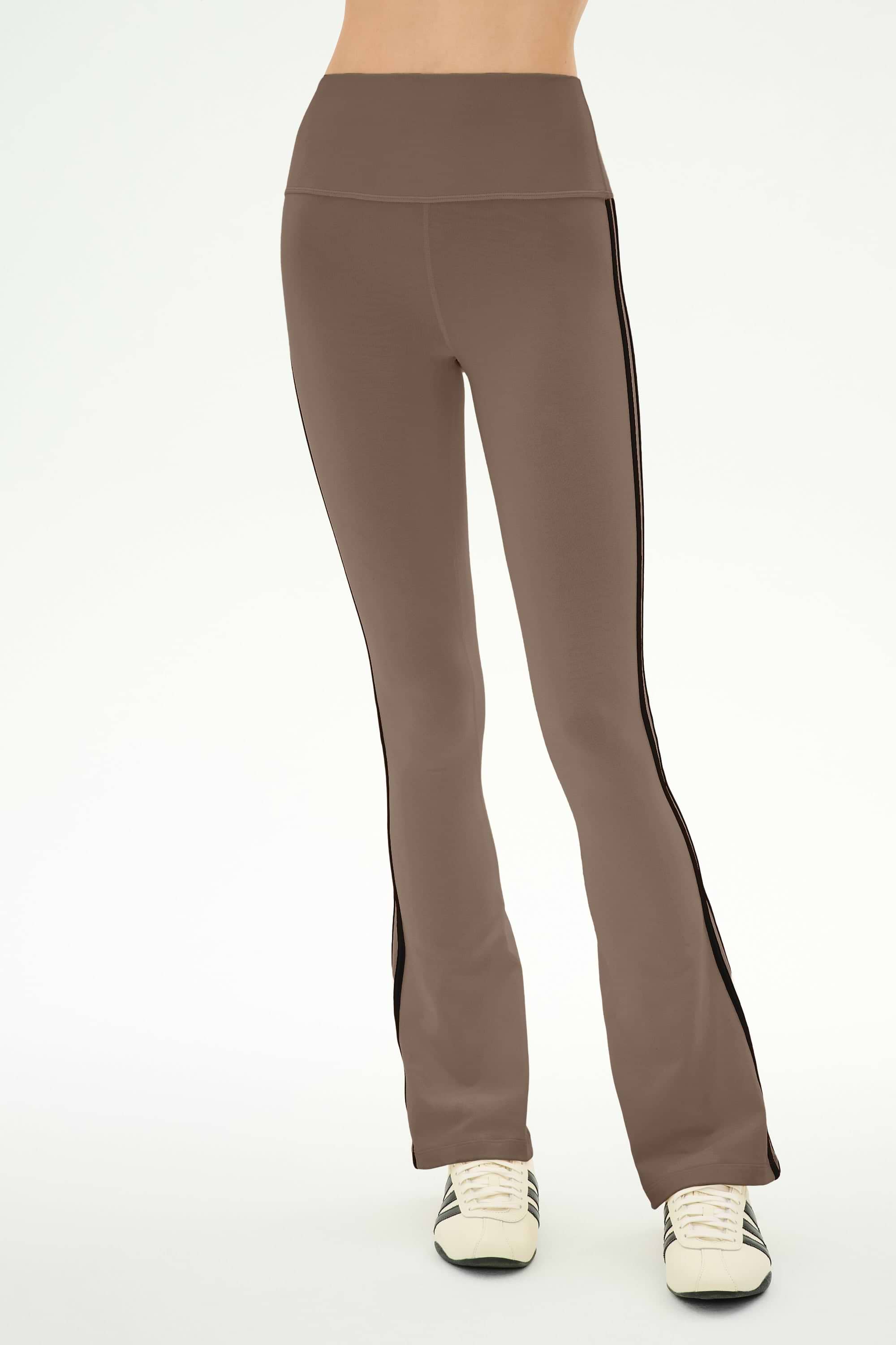 A person wearing the SPLITS59 Raquel High Waist Supplex Flare pants in Lentil/Black—a taupe, high-waisted flared style featuring black side stripes—paired with white sneakers, stands against a plain light background.