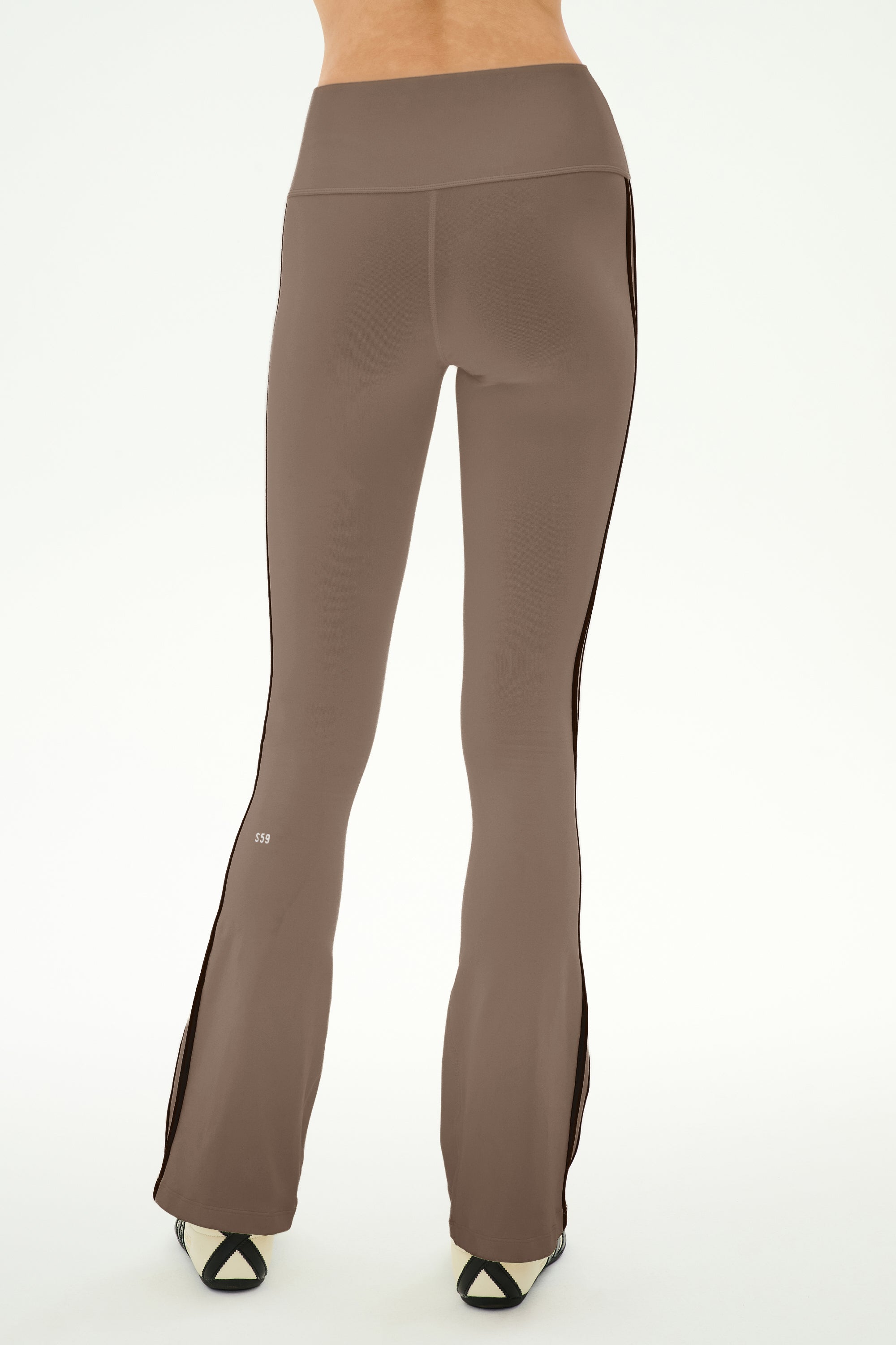 A person is seen from the back, standing on a white background, wearing the SPLITS59 Raquel High Waist Supplex Flare pants in Lentil/Black—a high-waisted brown flare style with black side stripes, crafted from a soft nylon-spandex blend.