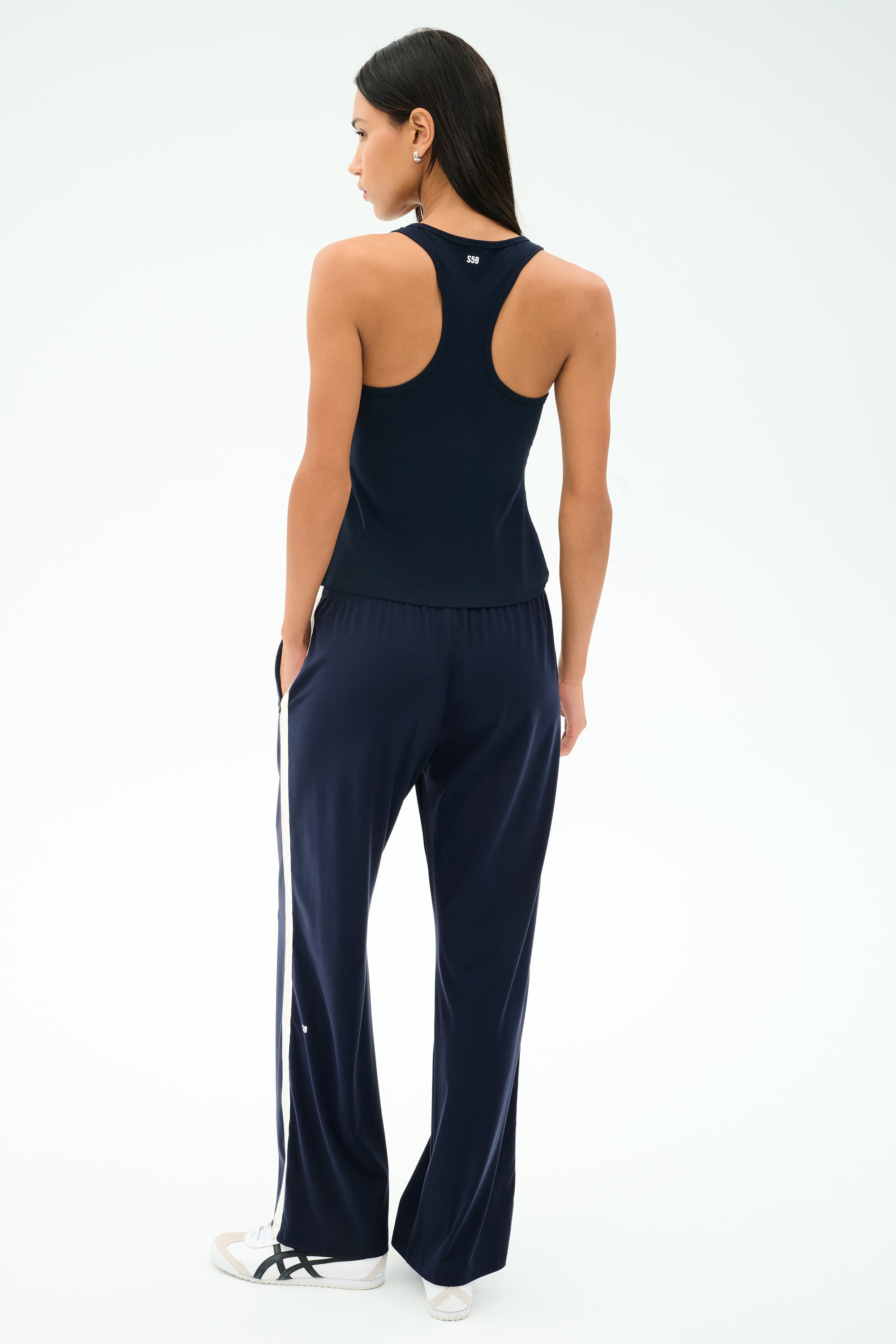 A woman faces away, wearing the SPLITS59 Mel Airweight Lite Tank in Indigo with matching wide-leg pants, both featuring white side stripes, paired with white sneakers.