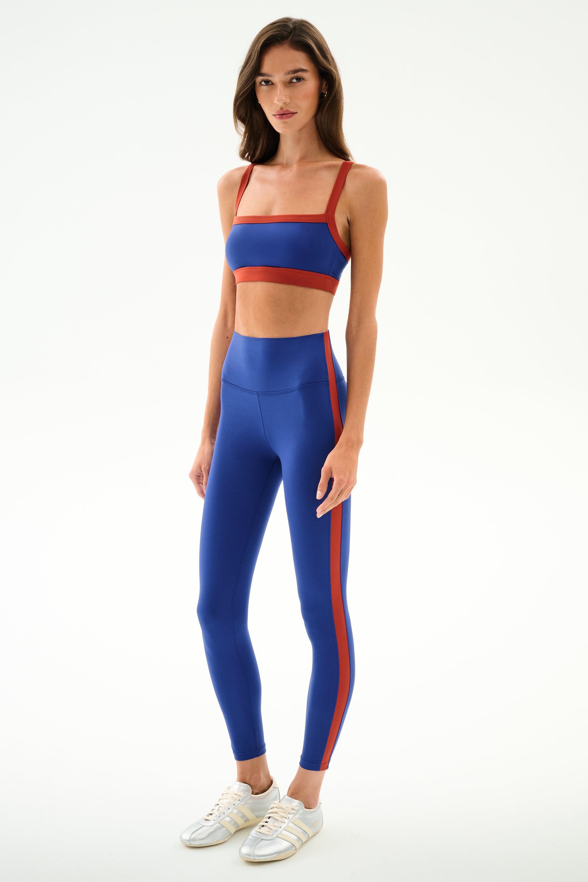 A woman models the SPLITS59 Monah Rigor Bra - Cobalt/Terracotta with matching leggings and white sneakers in a studio with a plain white background.