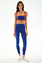 A woman wears the SPLITS59 Clare High Waist Rigor 7/8 - Cobalt/Terracotta leggings set with white sneakers, standing against a plain white background.