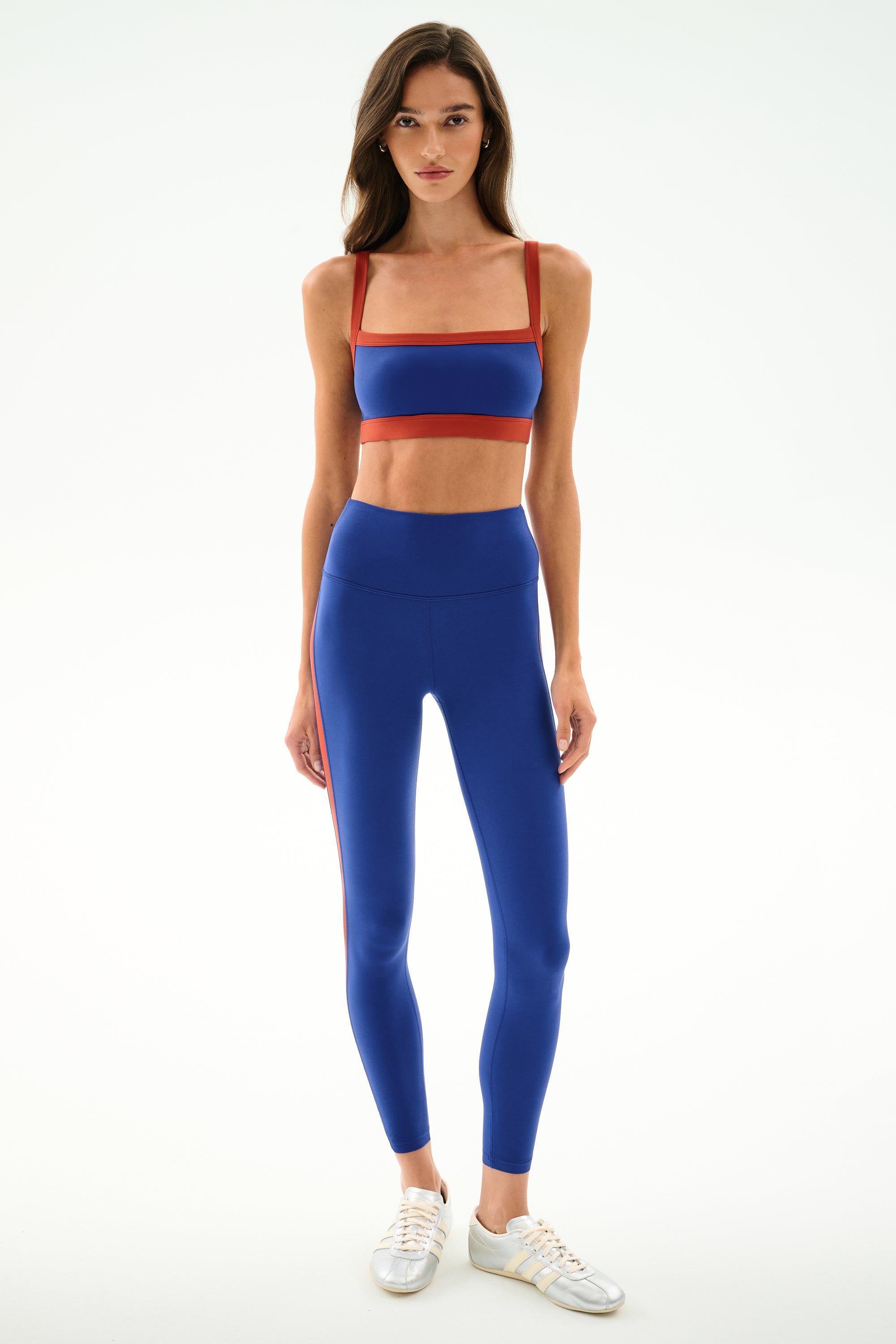 Woman wearing the SPLITS59 Monah Rigor Bra - Cobalt/Terracotta with matching leggings and white sneakers, standing against a plain white background.