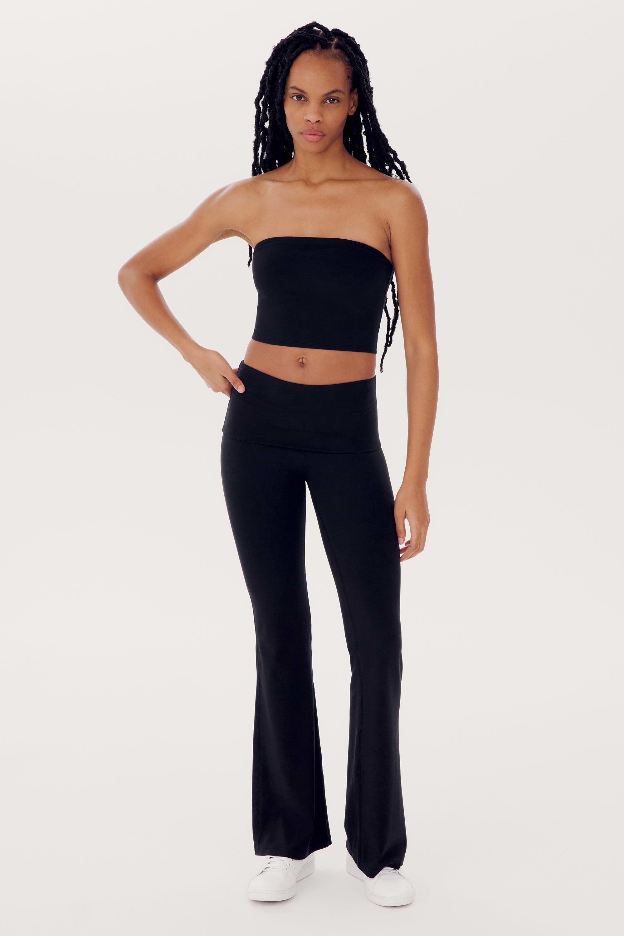 A person with long hair in a long braid stands against a plain background wearing the SPLITS59 Hazel Airweight Crop Bandeau - Black, black flared spandex pants, and white shoes.