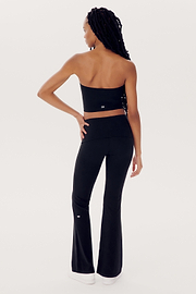 A person with long braids is standing with their back to the camera, wearing a strapless black top (Hazel Airweight Crop Bandeau - Black by SPLITS59), black flared pants made of spandex, and white sneakers against a plain white background.