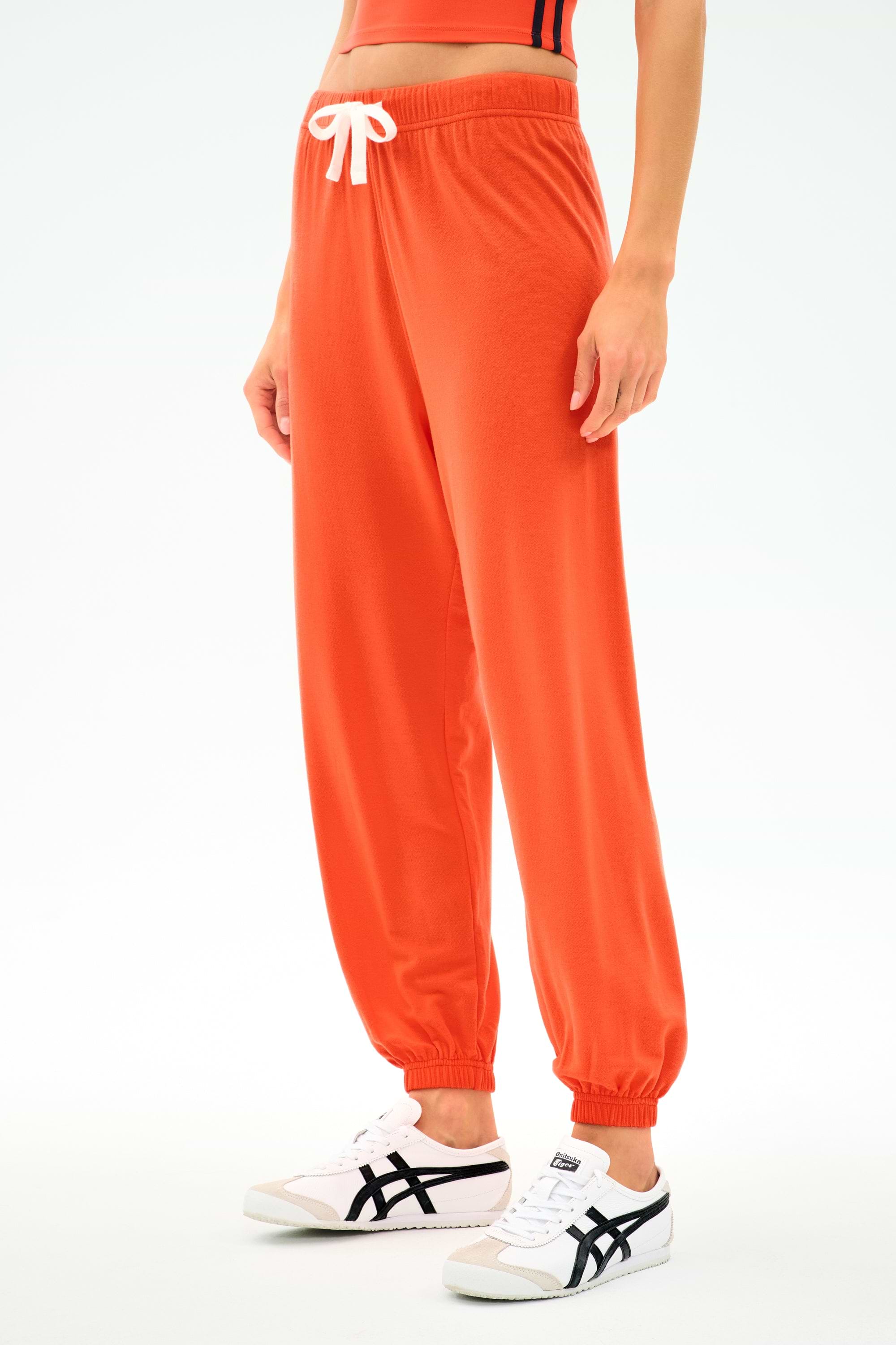A person wearing SPLITS59's Andie Oversized Fleece Sweatpant in Valencia, an eye-catching orange, paired with white sneakers with black accents, stands against a plain light background.