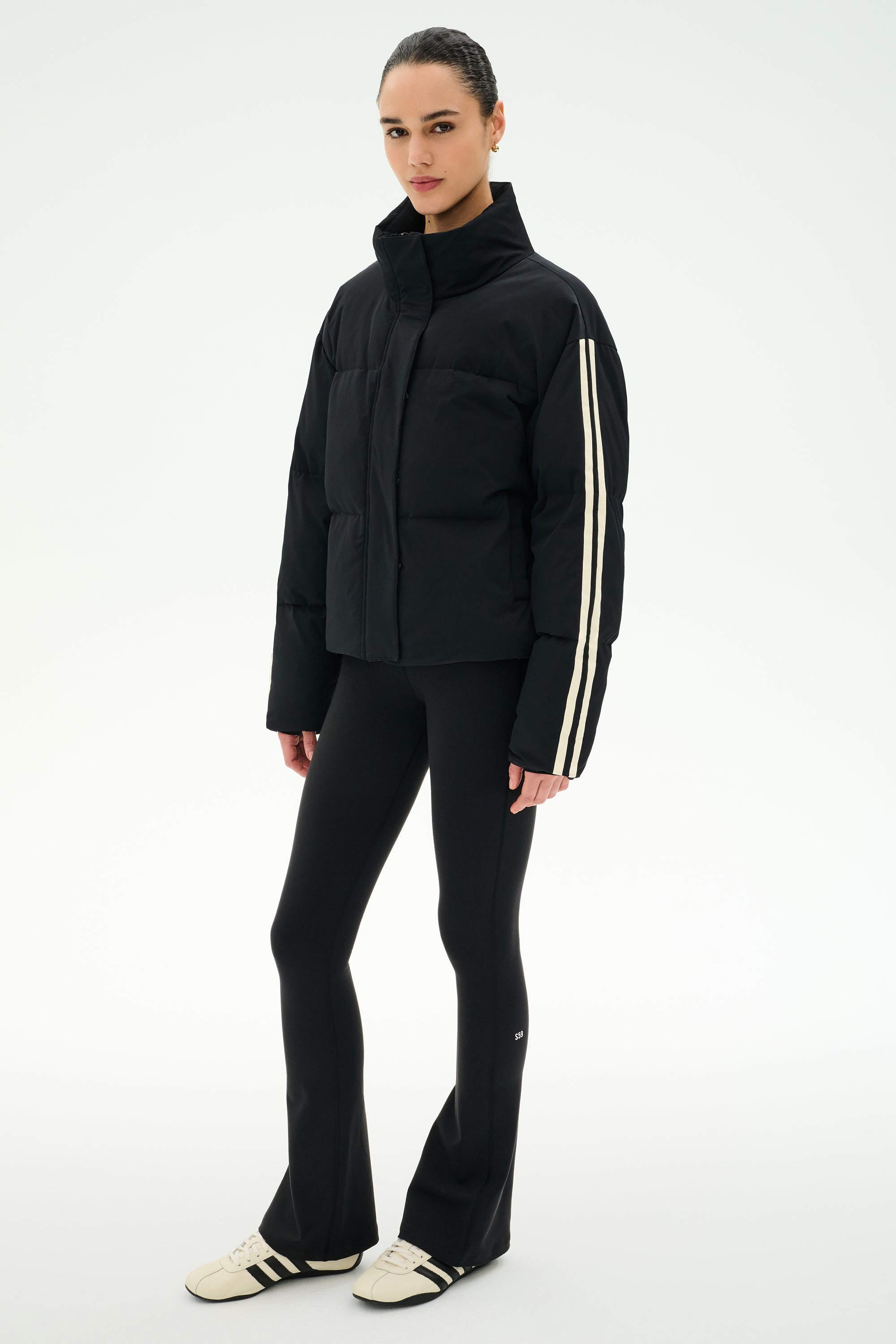 A person wearing the SPLITS59 Maeve Puffer Jacket in Black/Creme—crafted from 100% nylon with distinctive white sleeve stripes—paired with black flared pants and white sneakers, stands against a plain white background.