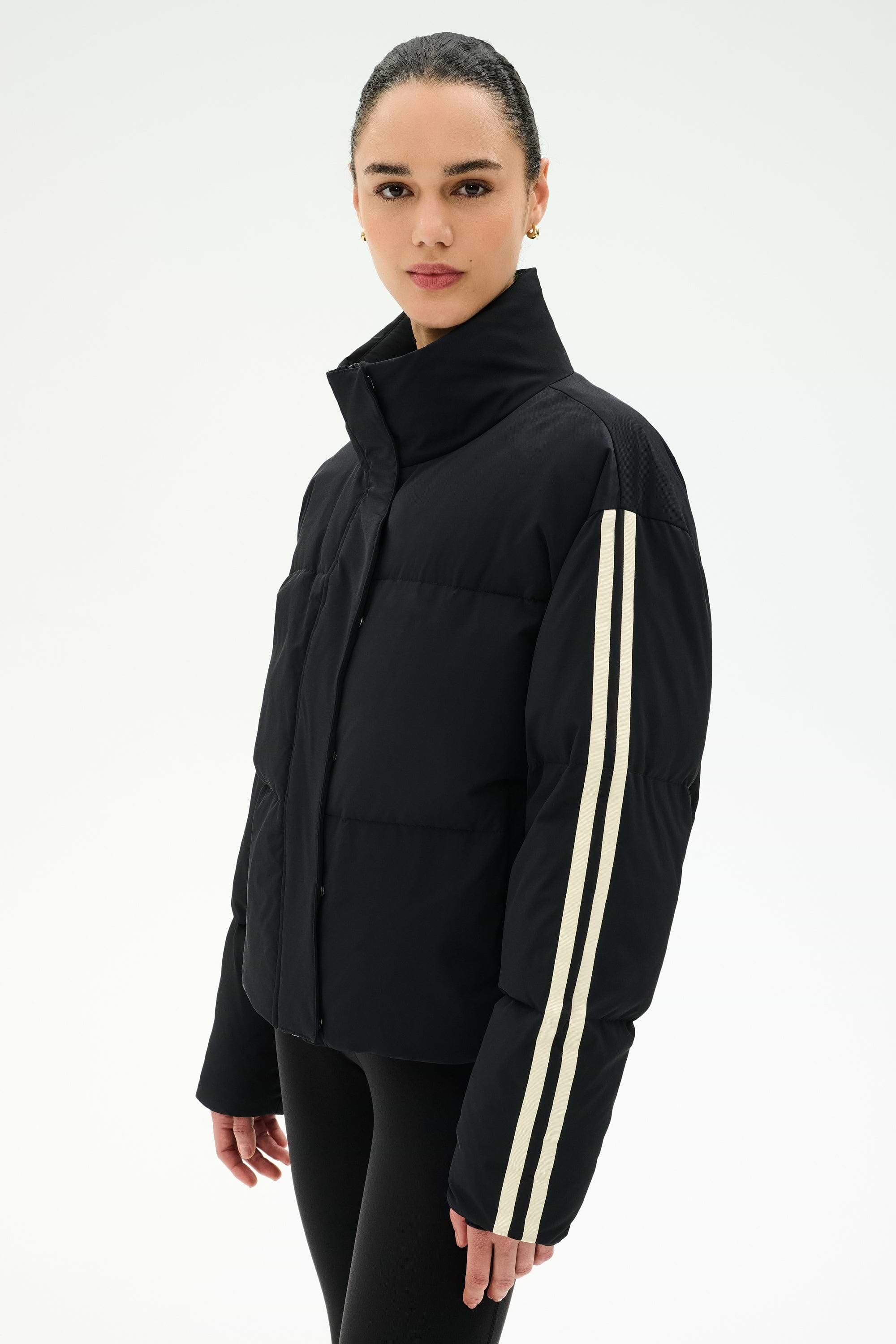 A woman models the SPLITS59 Maeve Puffer Jacket in Black/Creme, a 100% nylon jacket distinguished by two white stripes on the sleeve, standing against a plain light background.